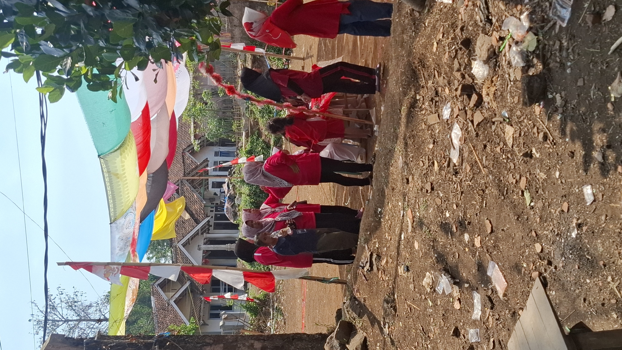 In the city of Cilaku, Indonesia, a group of individuals, predominantly women and children, are gathered in a dirt-covered outdoor area. Many are wearing bright red shirts and headscarves, suggesting a coordinated event or celebration. Some of the women appear to be interacting, with one holding a trophy or award. Colorful fabric banners, including Indonesian flags, are strung across the area, indicating a festive occasion. Behind them, residential buildings with tile roofs are visible, suggesting a semi-urban or village setting. The ground is uneven and strewn with debris, including plastic packaging and broken bricks, hinting at an undeveloped or rural environment. The sky is clear and blue, and the overall scene suggests a daytime event taking place under bright sunlight.