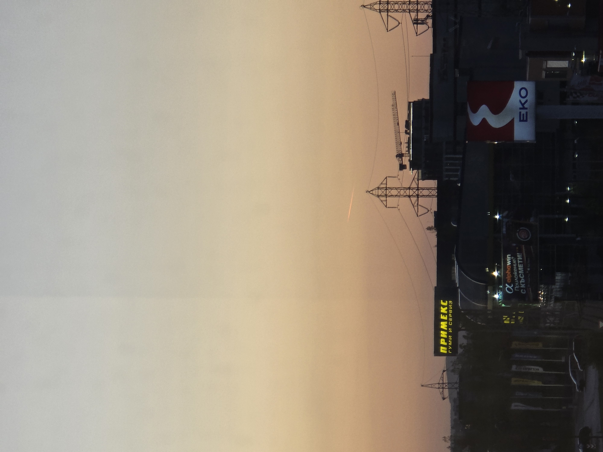 This image shows a dusk urban scene, likely in Sofia, Bulgaria, given the presence of several Cyrillic-script signs and brands specific to the area. Visible at dusk the sky transitions from light to dark on the left. High-voltage power lines are prominent, with their support towers spanning the composition. The silhouette of buildings and billboards is discernible against the sky. The "EKO" logo and Cyrillic script signs appear to be advertising various commercial entities, suggesting the presence of commercial areas in the city. In the foreground, dark silhouettes hint at vehicles and urban infrastructure.