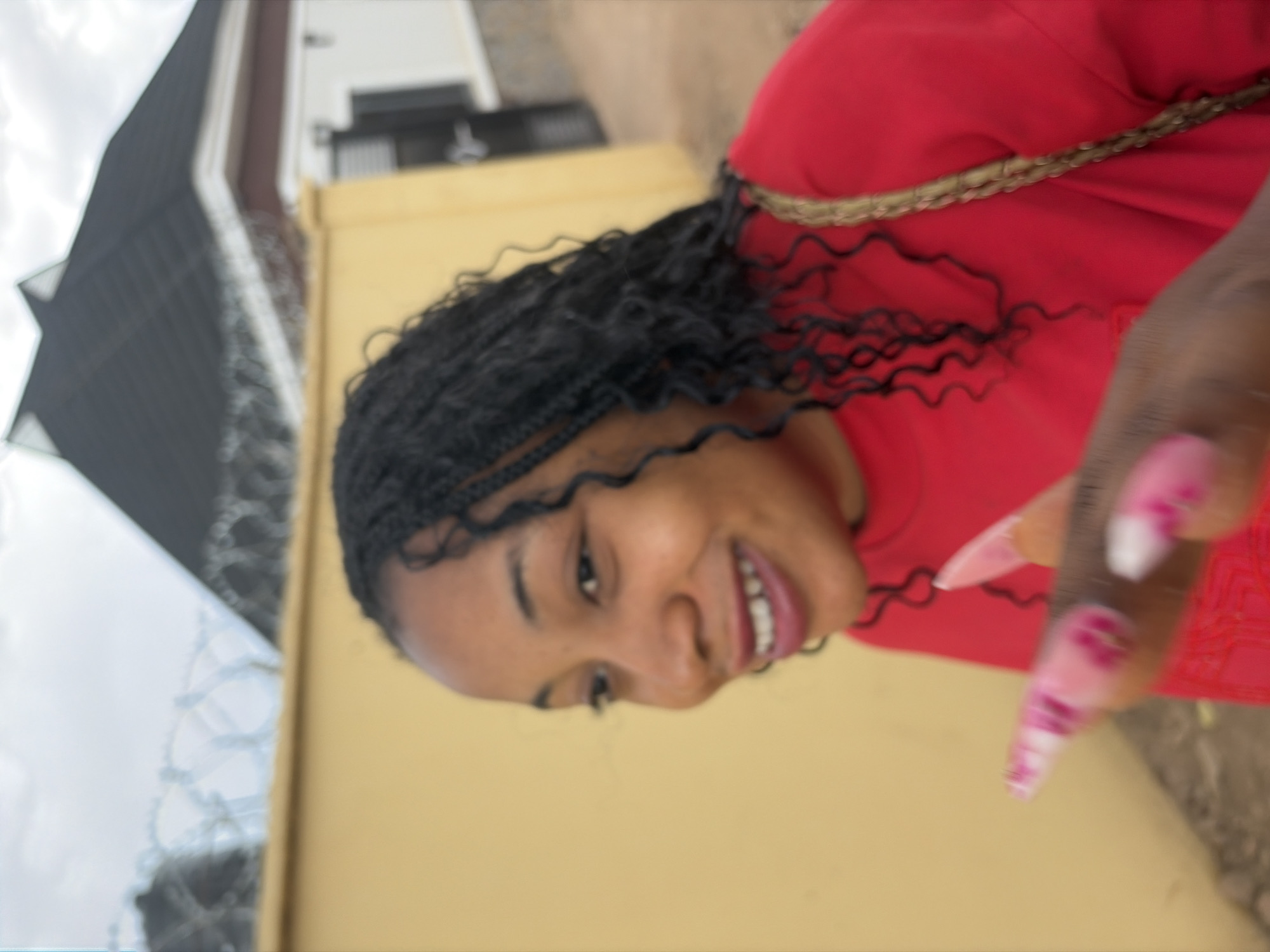 A woman with dark braided hair is wearing a red top with a gold braided trim. She is smiling and has long, pink and white painted fingernails. The background is a light yellow wall. A building with a dark roof and wire fencing is partially visible on the left side of the image. The location is Jos, Nigeria, based on visual cues.