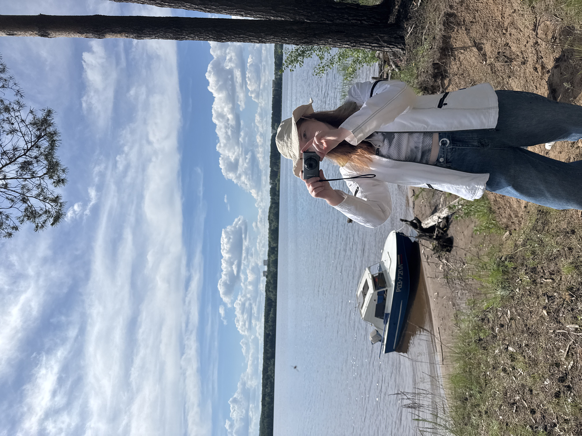 This image depicts a person standing on the bank of a wide body of water, likely a river or large lake, in a natural setting near Chaikovsky, Russia. The individual, a young female with reddish-brown hair, is actively engaged in photography, holding a black Canon compact digital camera up to her face, seemingly framing a shot. She is dressed in a white jacket, blue jeans, and a light-colored bucket hat.
The scene is set during the daytime under a bright sky filled with both fluffy cumulus clouds and high, wispy cirrus clouds. The expansive water surface stretches into the distance, meeting a distant, densely forested shoreline. The immediate bank is a mix of sandy earth and dirt, with patches of sparse grass and some visible roots or driftwood. Parts of tall pine trees frame the composition on both sides, indicating a surrounding forest.
To the right of the person, a small blue and white motorboat is partially beached on the shore. The text "РОЗ-720И" is clearly visible on the side of its hull. The image conveys a sense of peaceful outdoor activity, capturing a moment of appreciation for the natural landscape.
