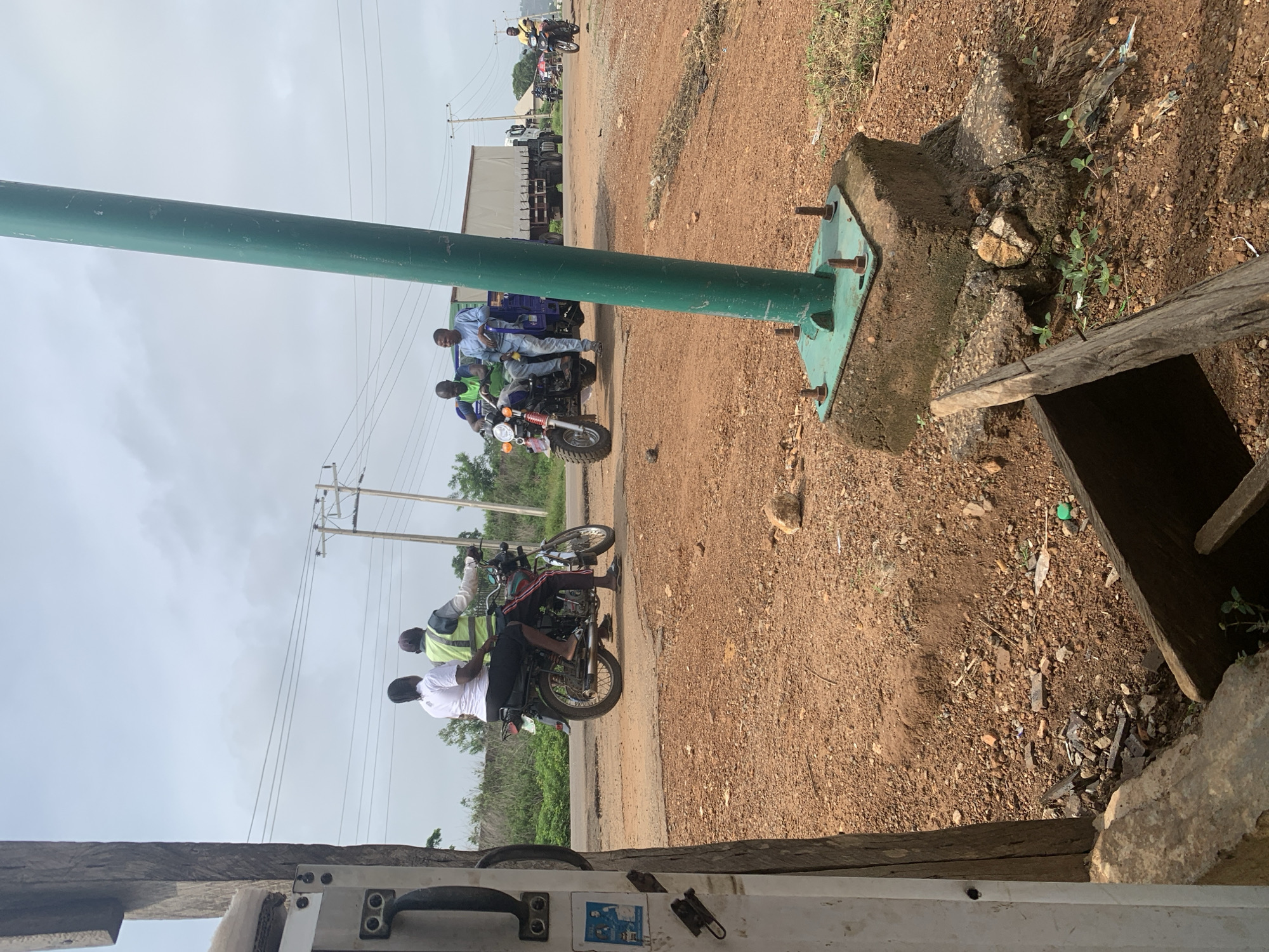 The original image is captured with a 90-degree clockwise rotation, depicting a roadside scene in what appears to be a semi-urban or rural setting. A large, thick green metal pole extends prominently across the frame, appearing horizontally due to the rotation. Its base, a square metal plate secured by bolts, is embedded in a concrete foundation on the reddish-brown, dusty ground. To the right of the pole's base, a wooden plank extends over a dark, recessed opening, possibly a ditch or drain, surrounded by scattered debris and sparse vegetation.
On a dirt road to the left of the pole, several men are gathered on motorcycles. One man, dressed in a light blue shirt and trousers, sits on a blue three-wheeled motorcycle or tricycle. Behind him, another man in a green safety vest and dark shirt is on a standard motorcycle, with at least two other men also on motorcycles further down the road. Power lines stretch across the overcast sky in the background. Beyond the road, large trucks or trailers are visible, along with some distant foliage. The scene appears to take place during the daytime under a hazy or overcast sky. No clearly legible text is visible in the image.