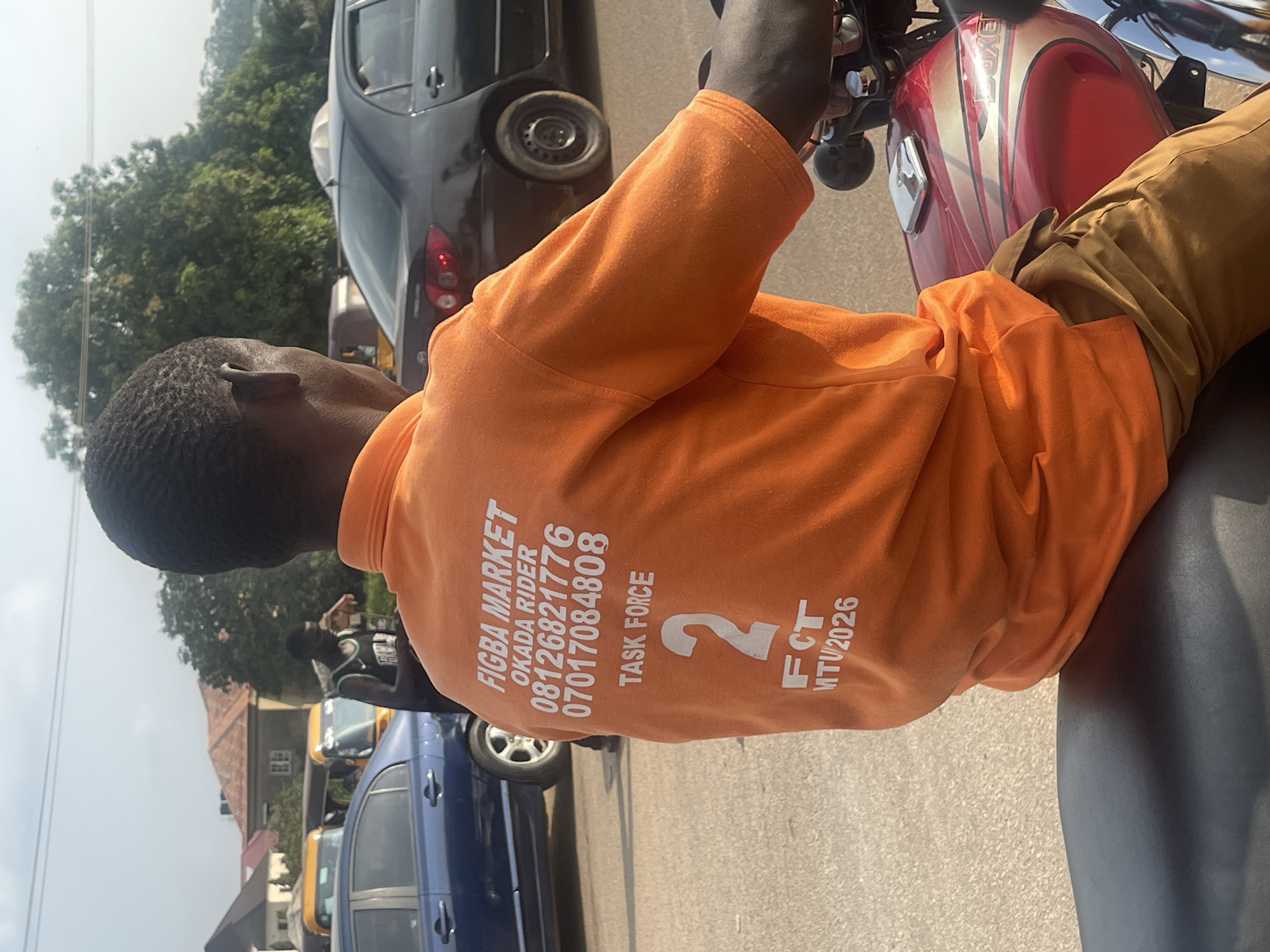 The image shows a back view of a person sitting on a red and gold motorcycle in a city street. The person is wearing an orange t-shirt with white text that reads "FIGBA MARKET OKADA RIDER 08126821776 07017084808 TASK FORCE 2 FCT MTV/2026". They are wearing brown pants. The motorcycle's fuel tank is visible, and a chrome component is on top of it. The background shows a street with several vehicles, including a blue car, a black car, and yellow tricycles, commonly known as "keke napeps" in Nigeria. Buildings and trees are also visible in the background, suggesting an urban or semi-urban setting. The sky is clear, indicating daylight. The scene appears to be in Galadimawa Village, Nigeria, based on the presence of okada riders, keke napeps, and the general streetscape.