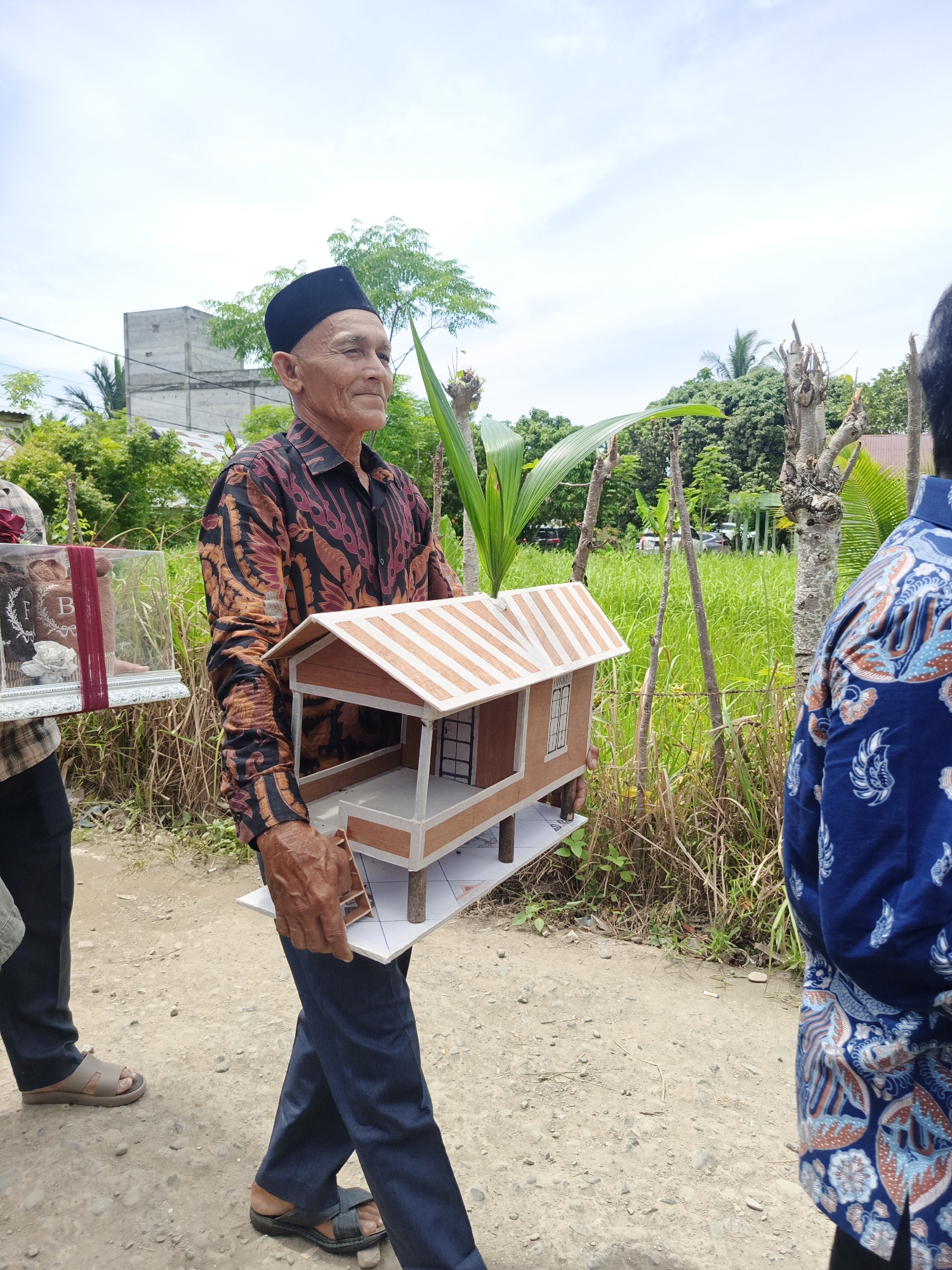 An elderly Indonesian man wearing a black peci hat and a batik shirt walks along a dirt path, carrying a miniature wooden house. He is accompanied by at least two other individuals, one of whom is visible from the back wearing a blue batik shirt. The environment appears to be a rural or semi-rural area with lush green vegetation, trees, and a partially visible building in the background. The sky is cloudy. The man's expression is calm as he looks to his right. The scene is likely taking place outdoors, possibly during a procession or a cultural event. The location in the city of Meuliek, Indonesia, is suggested by the context provided.