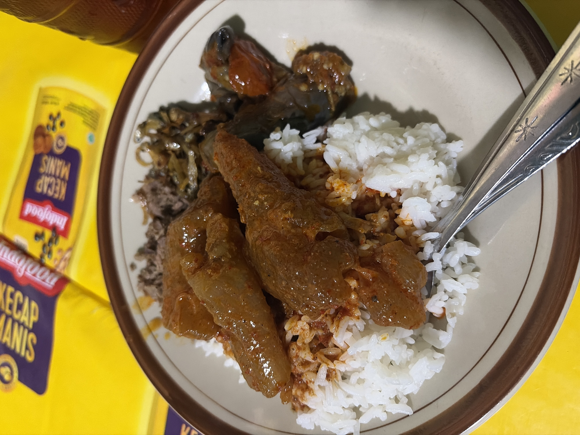 In Pekalongan, Indonesia, a plate of rice with Indonesian cuisine is presented on a table. The plate contains white rice topped with several pieces of what appears to be braised meat or offal in a reddish-brown sauce, along with some fried anchovies and perhaps vegetables. A fork rests on the side of the plate. In the background, a yellow packaging of "Indofood Kecap Manis" (sweet soy sauce) is visible, suggesting its use as an ingredient or condiment. A glass of iced tea is also partially visible to the right. The overall scene depicts a casual meal setting.