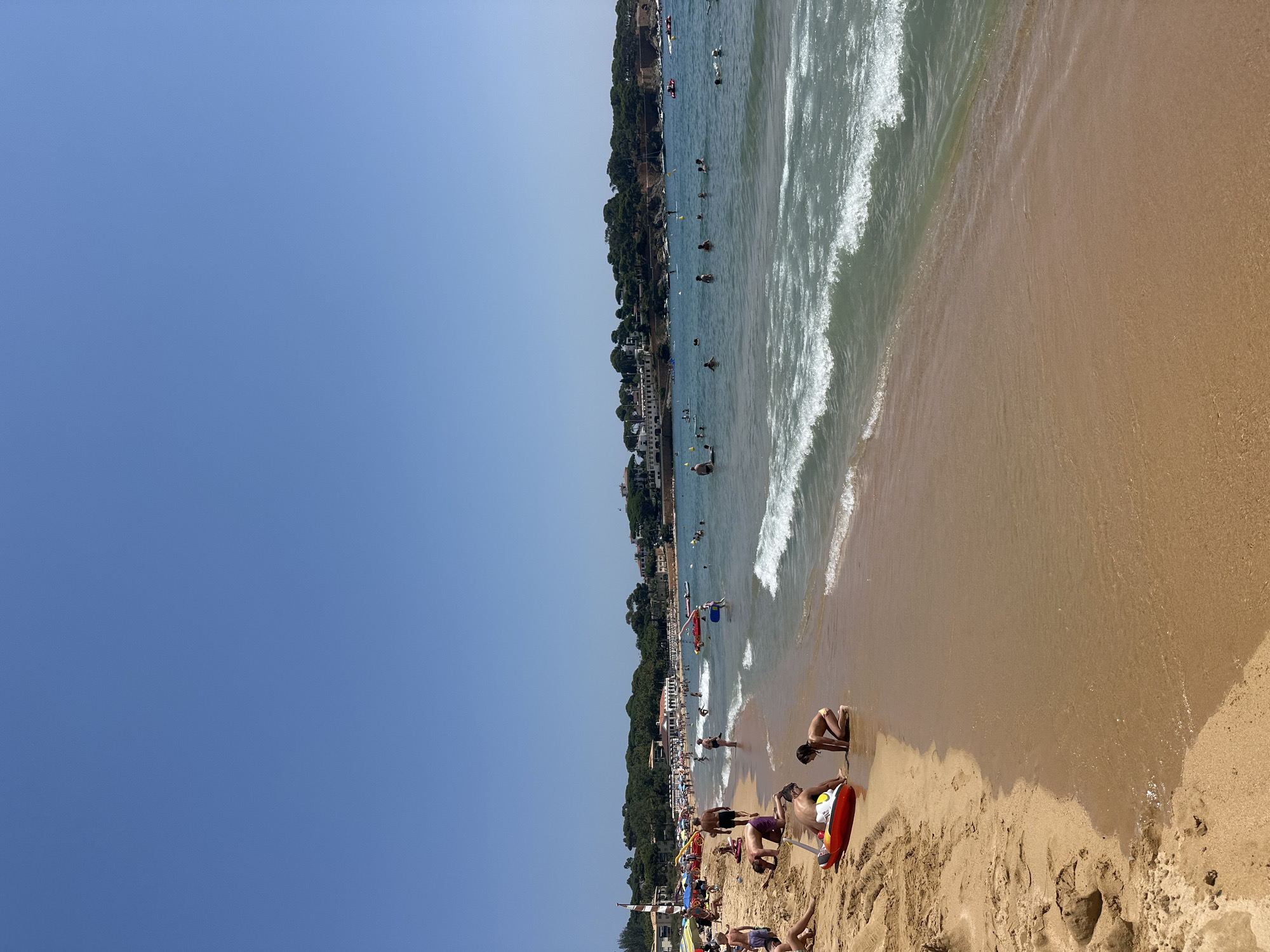 The image depicts a busy beach scene in Sant Feliu de Guíxols, Spain, on a clear, sunny day. The foreground is dominated by a sandy beach with several individuals engaged in various activities. Closer to the water, two children are playing with a red inflatable toy. Further up the beach, a man and a woman are visible, with the man wearing purple shorts and the woman in a bikini. Several other people are scattered across the sand, some lying down and others sitting.
The shoreline is lined with gentle waves breaking on the sand. The water is populated with numerous swimmers and a few people standing on paddleboards. In the background, a stretch of coastline is visible, featuring buildings, lush green trees, and fortifications. The sky above is a clear, vibrant blue, typical of a summer day. The scene conveys a lively atmosphere of a popular tourist destination.