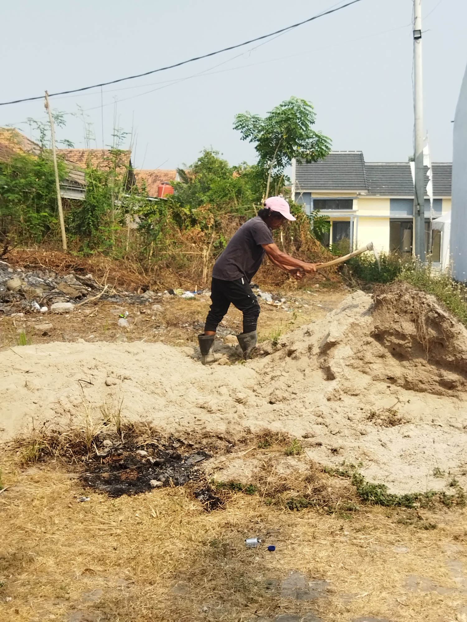 The image depicts an outdoor scene in Suka Sari, Indonesia, focusing on a man engaged in manual labor. The man, wearing a dark t-shirt, dark pants, black boots, and a pink cap, is positioned in the center of the frame. He is actively using a pickaxe to break apart a large pile of light-colored soil or sand. His posture suggests exertion as he swings the tool.
The environment appears to be a construction or land preparation site. In the background, partially obscured by foliage, are residential buildings with tiled roofs, characteristic of Indonesian architecture. Some of the buildings have yellow and gray exteriors. There is a significant amount of dry vegetation, including grasses and branches, scattered throughout the foreground and surrounding the soil pile. Some areas show signs of burnt material, indicating a possible past fire. A tall utility pole with a light fixture is visible on the right side of the frame, and electrical wires stretch across the upper portion of the image. The sky is clear and pale blue, suggesting daytime with bright sunlight. The overall impression is one of ongoing work in a semi-developed or developing area.