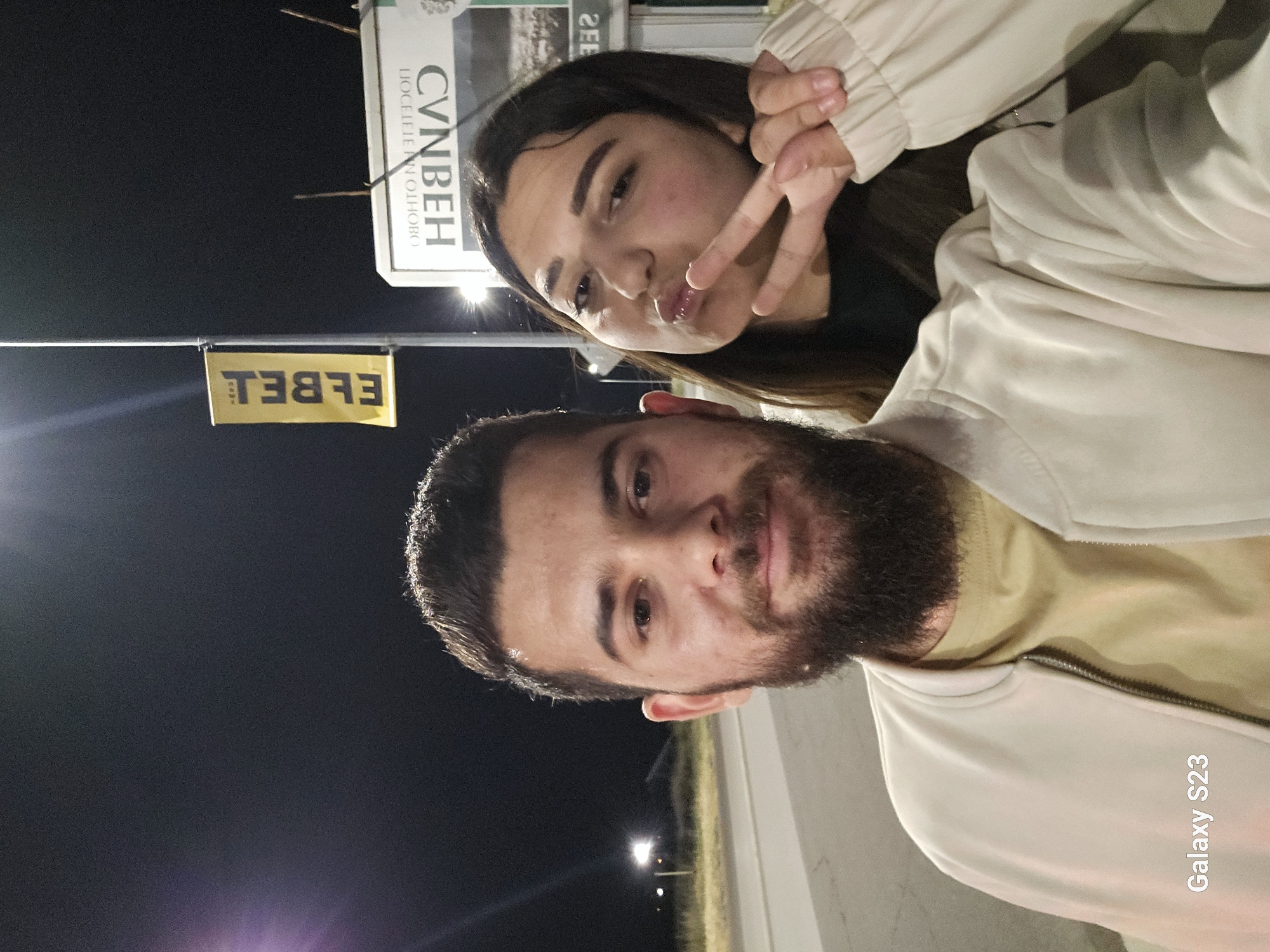 A selfie featuring a man and a woman, taken at night in Sliven, Bulgaria. The man has a dark beard and is wearing a light-colored jacket over a tan shirt. The woman has long brown hair and is making a peace sign with her fingers. Behind them, a sign with Cyrillic writing and another sign with "Elbel" written on it are visible. The background is dark, suggesting a nighttime setting. The photo was taken with a Samsung Galaxy S23.