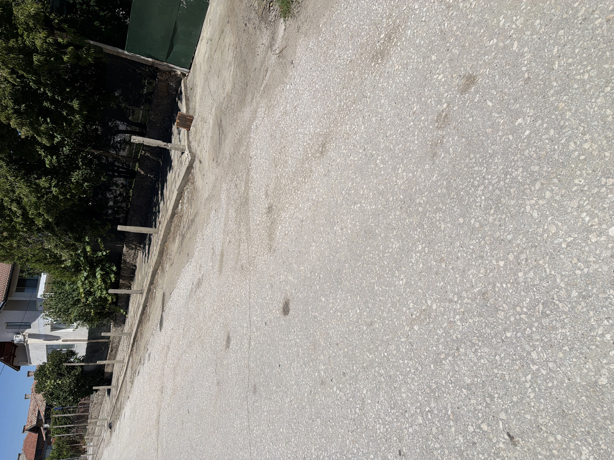 This is a ground-level, eye-level, wide-angle, outdoor, daytime photograph of a street in the village of Zlatitrap, Bulgaria. The street is paved with asphalt and shows some signs of wear, including darker patches that may be oil stains or water. A concrete sidewalk runs along the right side of the street, lined with numerous concrete posts, likely for traffic or pedestrian control. Behind the sidewalk, there is a low stone wall and then dense green foliage and trees. A white building with a balcony and an air conditioning unit is visible above the foliage, and further down the street, other buildings with tiled roofs can be seen. A large, dark green metal gate is present on the far right side of the image, and a small, reddish-brown wooden block lies on the ground near the gate. The sky is clear and blue. There are no people or animals visible in the frame.