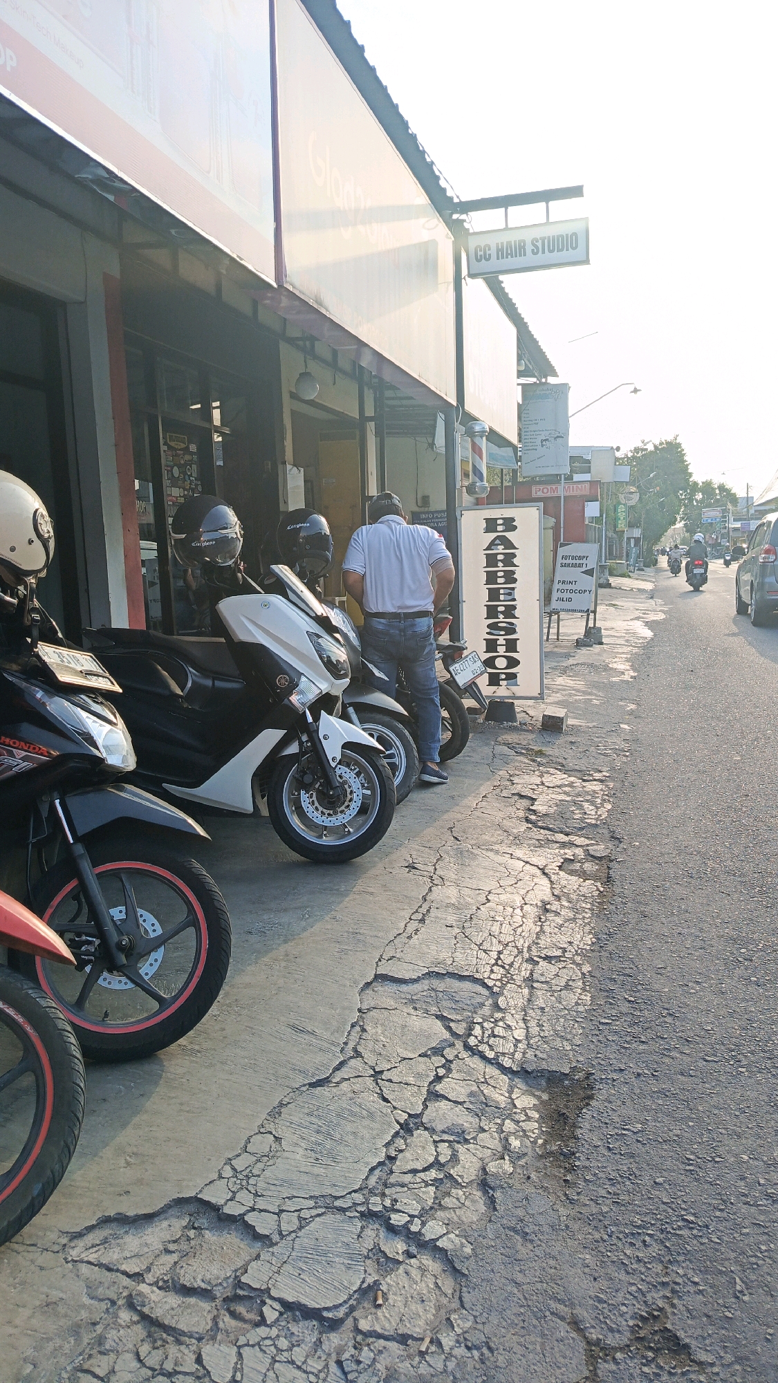 The image depicts a street scene in Ronowijayan, Indonesia, with a focus on a barbershop and surrounding parked motorcycles. A man in a white shirt and blue jeans stands facing away from the camera, near a white scooter. To his left, several motorcycles are parked along the sidewalk, including a black Honda with red accents and a white scooter. Above the shop, a sign reads "CC HAIR STUDIO." A vertical sign with "BARBERSHOP" printed in large black letters stands on the sidewalk. The sidewalk itself is cracked and uneven, transitioning to a paved road where traffic, including motorcycles and a car, is visible in the distance. The scene is illuminated by the warm glow of sunlight, suggesting it is late afternoon.