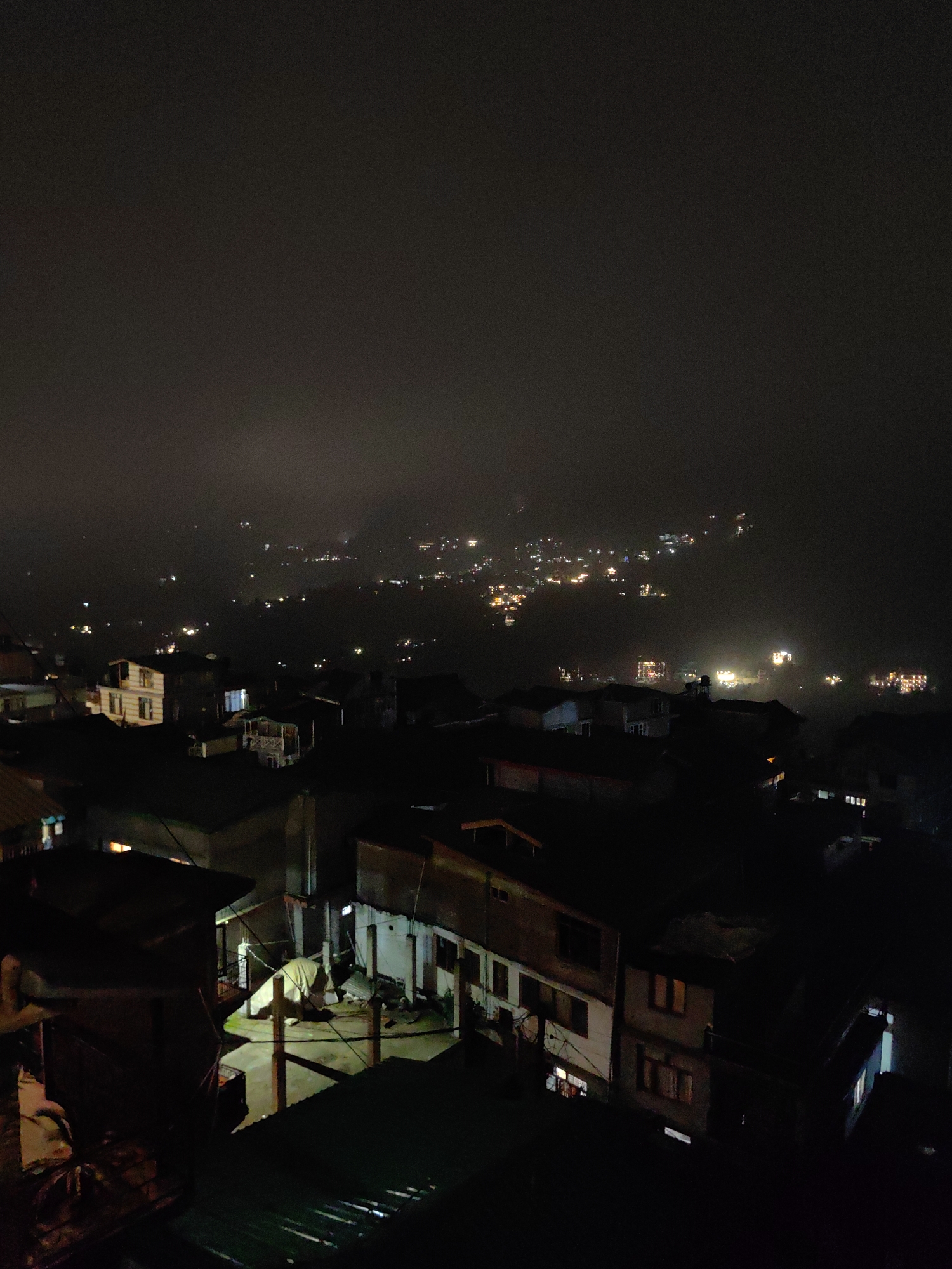 This is a nighttime aerial view of a densely populated urban area, likely a hillside community in Vashisht, India, given the characteristic architecture and the prevalent fog or mist contributing to the diffused lighting. The scene is dominated by darkness, with the primary illumination coming from numerous artificial lights emanating from buildings and streetlamps, creating a bokeh effect in the distance due to atmospheric conditions.
In the foreground, several buildings are visible, some with visible windows lit from within. The architecture appears to be a mix of traditional and more modern structures, with tiled roofs and multi-story dwellings. Some structures have an unfinished or dilapidated appearance. One building in the mid-ground, a light-colored, multi-story structure, stands out with multiple lit windows. Further into the background, the town sprawls across a hilly terrain, with clusters of lights indicating more development on the slopes.
The atmosphere is hazy, and the fog obscures finer details, giving the distant lights a soft, glowing appearance. There are no visible people or overt actions depicted in the image. The overall impression is one of a quiet, dimly lit settlement at night, shrouded in mist. The presence of multiple structures, interconnected by what appear to be wires or cables, suggests a residential or mixed-use area. The scene is composed of dark shadows punctuated by scattered points of light, creating a mood of mystery and stillness.
