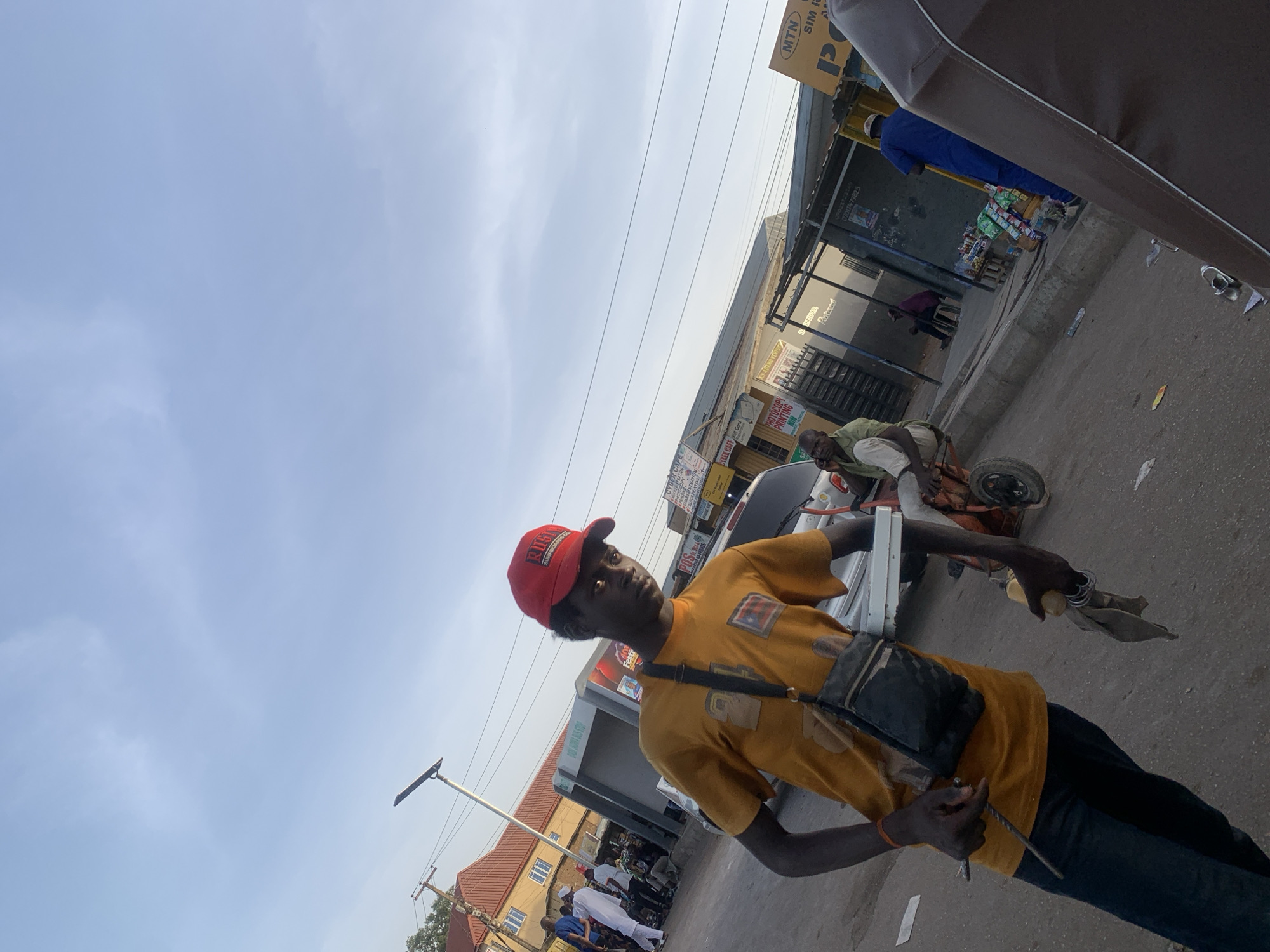 The image captures an urban scene in Jos, Nigeria. A young man wearing a red baseball cap and an orange t-shirt is the focal point. He has a black shoulder bag and is holding a metal rod and possibly cleaning materials. In the background, there's a street setting with buildings and shops displaying signage. Some visible signs are for MTN SIM cards and a cyber cafe offering services like photocopying and printing. People are present in the background, engaged in various activities on the street. A man is sitting on a small cart-like structure. The environment includes elements of urban infrastructure like a street lamp.