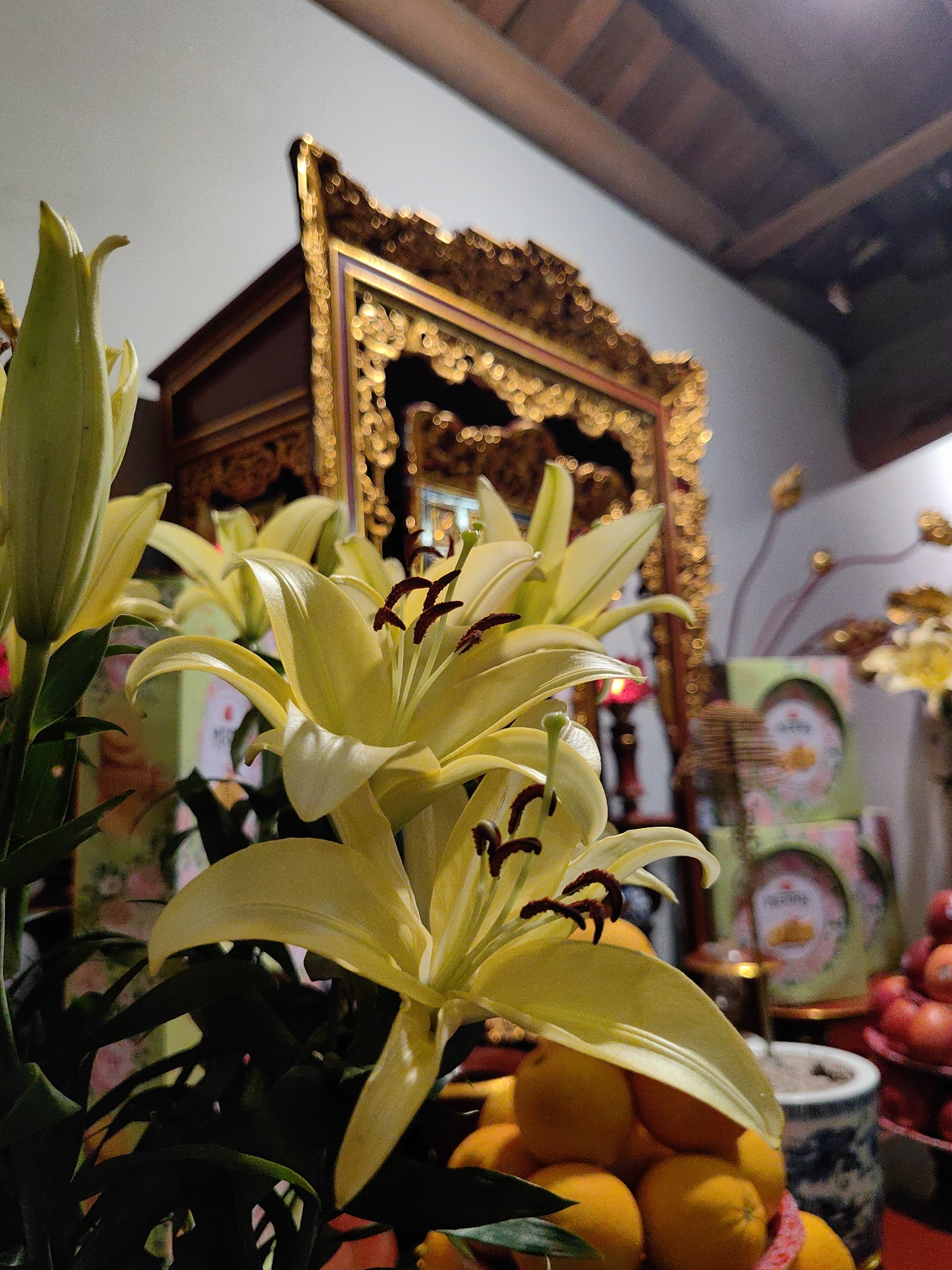 The image depicts a close-up of yellow lilies in front of a decorative, ornate gold-framed object, possibly a religious shrine or altar. A stack of gift boxes with circular designs and red and orange fruit are also visible in the background. The setting appears to be an interior space, likely a home. Based on the provided location of Hong Gai Ward, Vietnam, the scene likely represents a religious or cultural practice involving offerings of flowers and fruits.