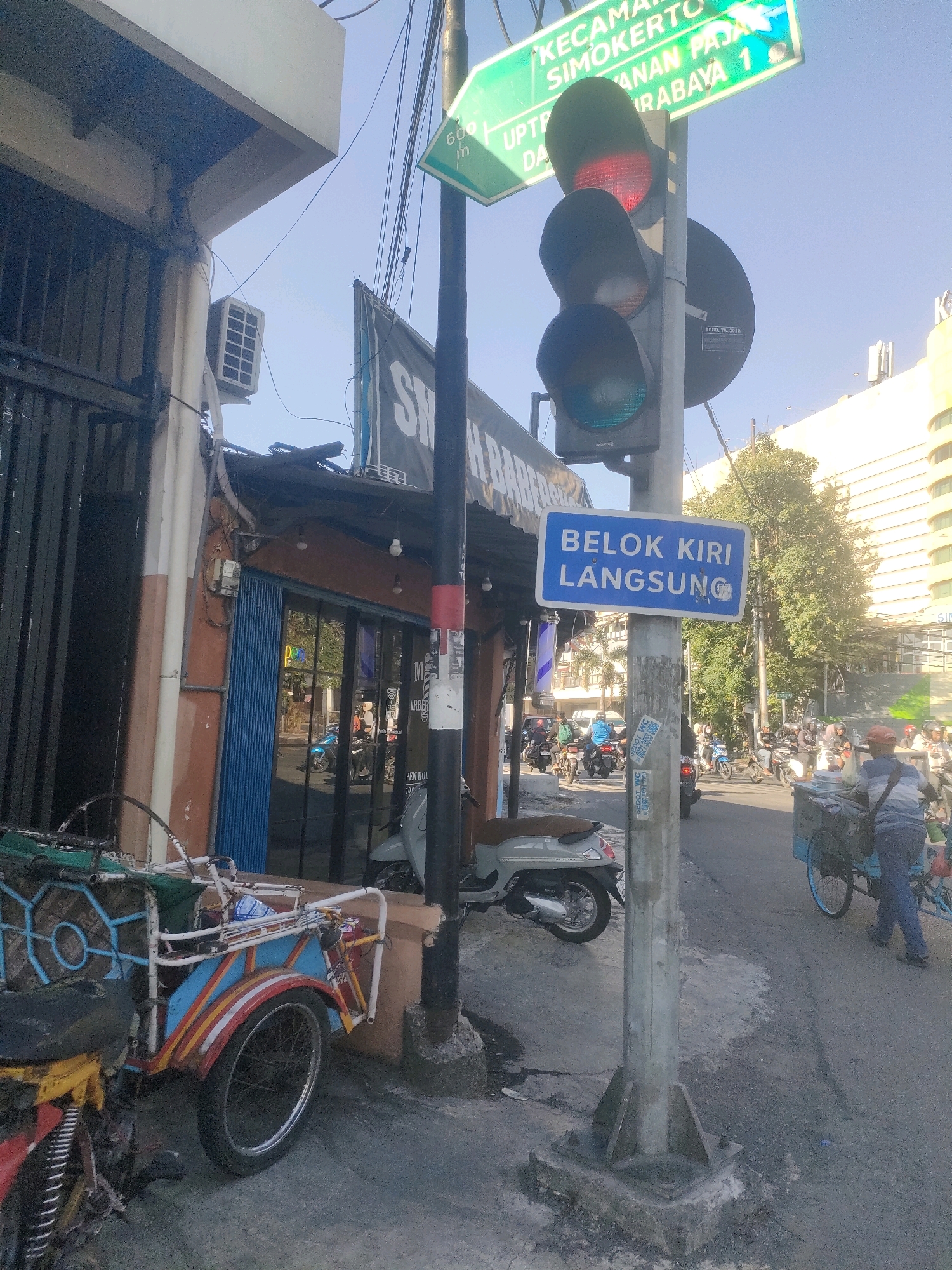 The image captures a street scene in Surabaya, Indonesia, specifically near the intersection indicated by street signs. A traffic light is prominently featured, with the red light illuminated. A blue sign below the traffic light reads "BELOK KIRI LANGSUNG," which translates to "TURN LEFT IMMEDIATELY."
To the left of the traffic signal, a building with an orange facade and a blue rolling door is visible, likely a commercial establishment. A sign above the entrance reads "SM..." indicating a business name. In the foreground on the left, a colorful, three-wheeled vehicle, possibly a becak or a modified motorcycle, is parked.
On the right side of the street, several motorcycles are parked and in motion, suggesting moderate traffic. A person is seen pushing a cart with a blue canopy. In the background, a tall, modern building with many windows is visible, contrasting with the older street-level establishments. The sky is clear and blue, indicating a sunny day.