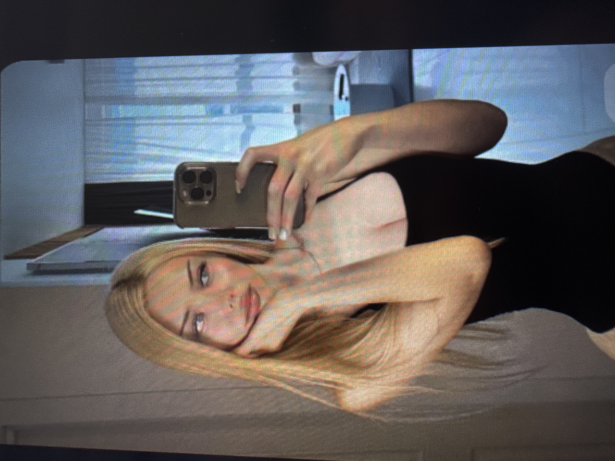 The image, rotated 90 degrees counter-clockwise, displays a close-up of a young blonde woman taking a mirror selfie. She has long, straight blonde hair that falls over her shoulders, light-colored eyes, and a fair complexion, with a slight, calm expression directed at the camera. She is wearing a fitted black top.
The woman holds a smartphone, identifiable by its prominent triple-camera array on the back, in her right hand to capture her reflection. Her left hand is positioned near her face, with fingers lightly touching her chin and neck.
The background, visible in the reflection behind her, suggests an indoor setting, possibly a bathroom, with light-colored walls and horizontal blinds or slats on a window. Faint outlines of what could be bathroom fixtures are also discernible in the background. The lighting appears to be indirect and even. The image exhibits a visible screen-door effect and pixelation, indicating it was likely captured as a photograph of a digital screen. No text is visible in the image.