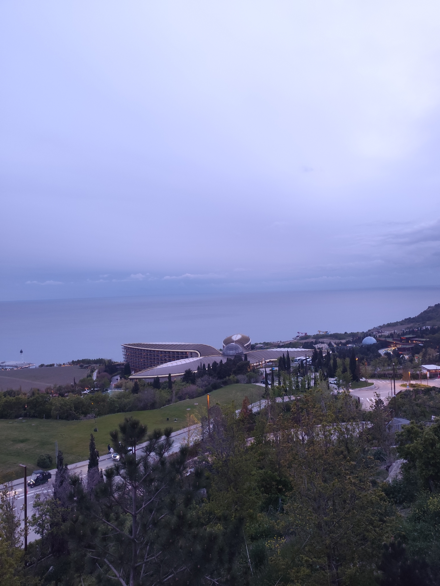 The image depicts a landscape in Симеизский поселковый совет, Ukraine. In the distance is the sea, with a cloudy sky above. A large, modern hotel complex sits on the coastline. The foreground contains a road with cars on it, surrounded by green trees and bushes. A few streetlights illuminate the scene. The overall light suggests it is either early morning or dusk.