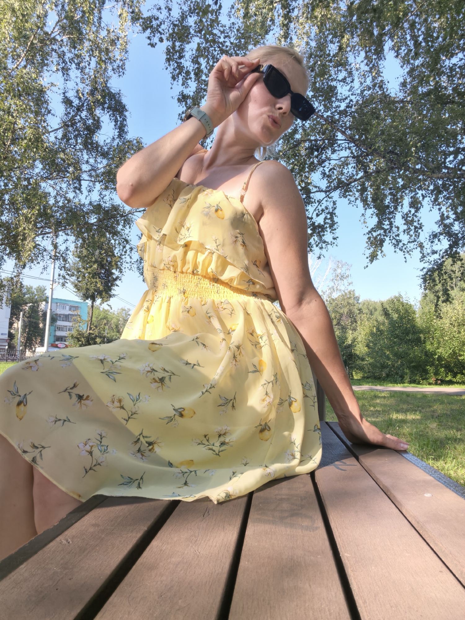 A blonde woman wearing a yellow floral print dress and black sunglasses sits on a wooden park bench, her left hand resting on the bench. She is looking towards the camera with her right hand raised, adjusting her sunglasses. The background shows trees, a clear blue sky, and some buildings in the distance. The image appears to have been taken outdoors during the daytime.