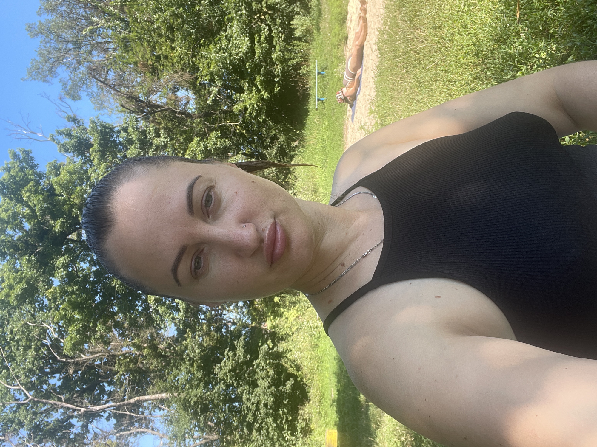 A woman with dark hair pulled back in a ponytail takes a selfie outdoors. She is wearing a black tank top and a silver necklace. Her expression is neutral, and she is looking directly at the camera. In the background, trees and a bright blue sky are visible. Further in the distance, a person in a striped bikini is lying on a towel on the sand, and a blue bench is present. The image appears to have been taken in a park or recreational area.
