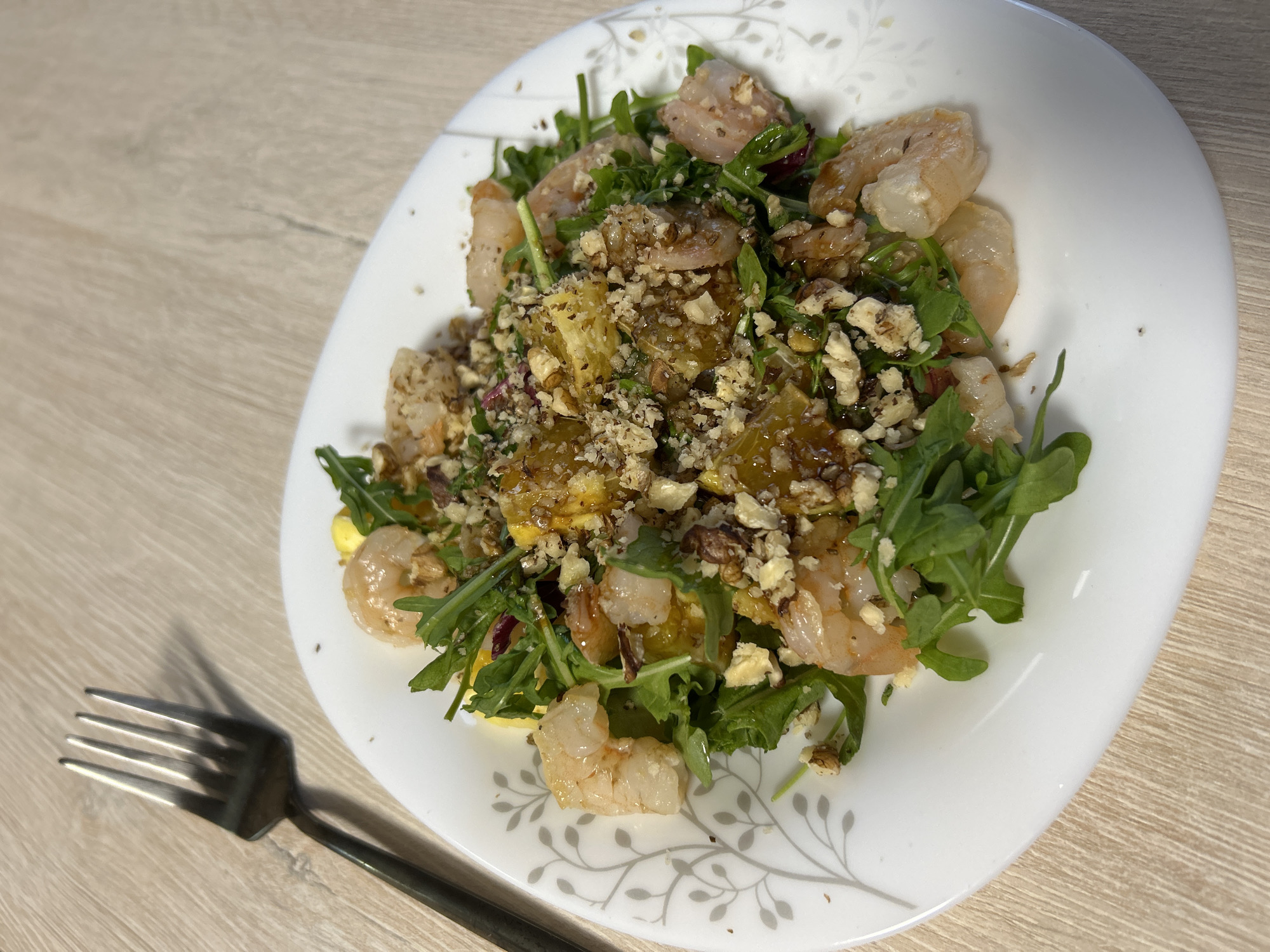 The image displays a vibrant salad served on a white ceramic plate, placed on a light-colored wooden surface. The main subject is a substantial and colorful salad, featuring a base of fresh green leafy vegetables, likely arugula or mixed greens. Mixed within the greens are several pieces of cooked, pinkish-white shrimp. Also visible are distinct chunks of a yellow-orange ingredient, which could be pineapple, mango, or squash. The entire dish is heavily garnished with finely chopped nuts, appearing to be walnuts or pecans, and a glossy, dark dressing, possibly a balsamic vinaigrette or glaze, is drizzled over the top.
The white plate has a subtle and elegant gray leaf and branch pattern decorating its rim. To the lower left of the plate, a dark-colored metal fork rests horizontally on the wooden surface, casting a clear shadow. The wooden surface exhibits a visible grain pattern. The lighting in the image is bright and even, indicating an indoor setting with good illumination, likely during the daytime. There are no people visible, and the activity depicted is the presentation of a prepared meal, ready for eating. No text is visible in the image. While the provided context suggests the location is Chaikovsky, Russia, there are no specific visual cues within the image itself that denote this geographical location.