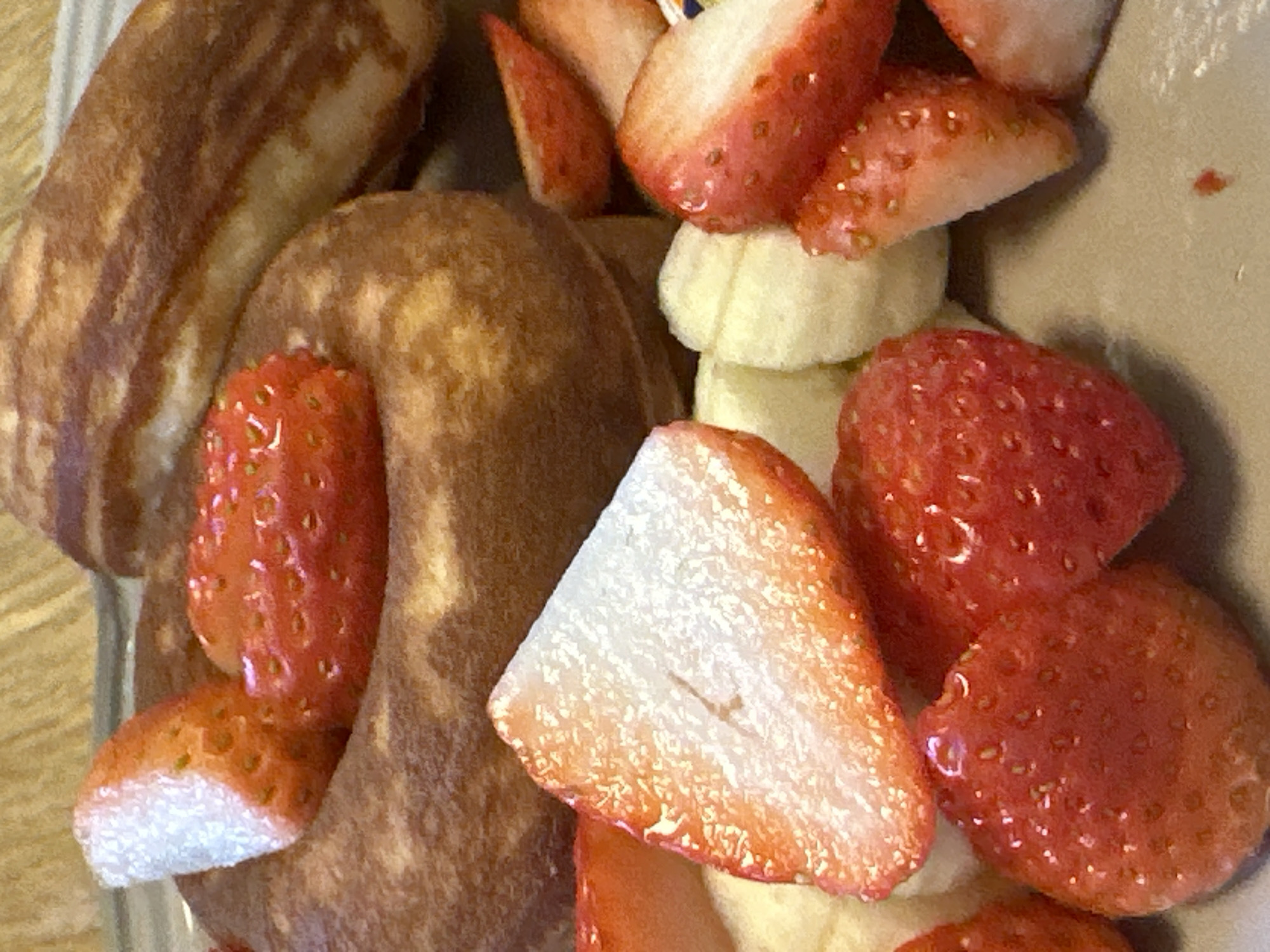 The image displays a close-up of food items arranged on a light-colored, flat surface, likely a plate. The central subject is a golden-brown, ring-shaped pastry, possibly a donut, with an irregular, textured surface. Resting within and around this pastry are numerous strawberries, some whole and bright red with visible seeds, others sliced in halves or quarters, exposing their white interior flesh.
In the upper right portion of the image, an elongated, cylindrical pastry with darker brown longitudinal stripes is partially visible, appearing to be a type of bread or sweet roll. Scattered among the strawberries and pastries are small, pale yellow or off-white chunks, consistent with sliced bananas or soft confectionary. The lighting is warm and direct, highlighting the textures and colors of the food.
No people are visible. The background is out of focus, presenting blurred warm tones, indicative of an indoor setting. There are no identifiable landmarks, signs, or environmental cues to determine a specific location within the city of Seoul, South Korea.