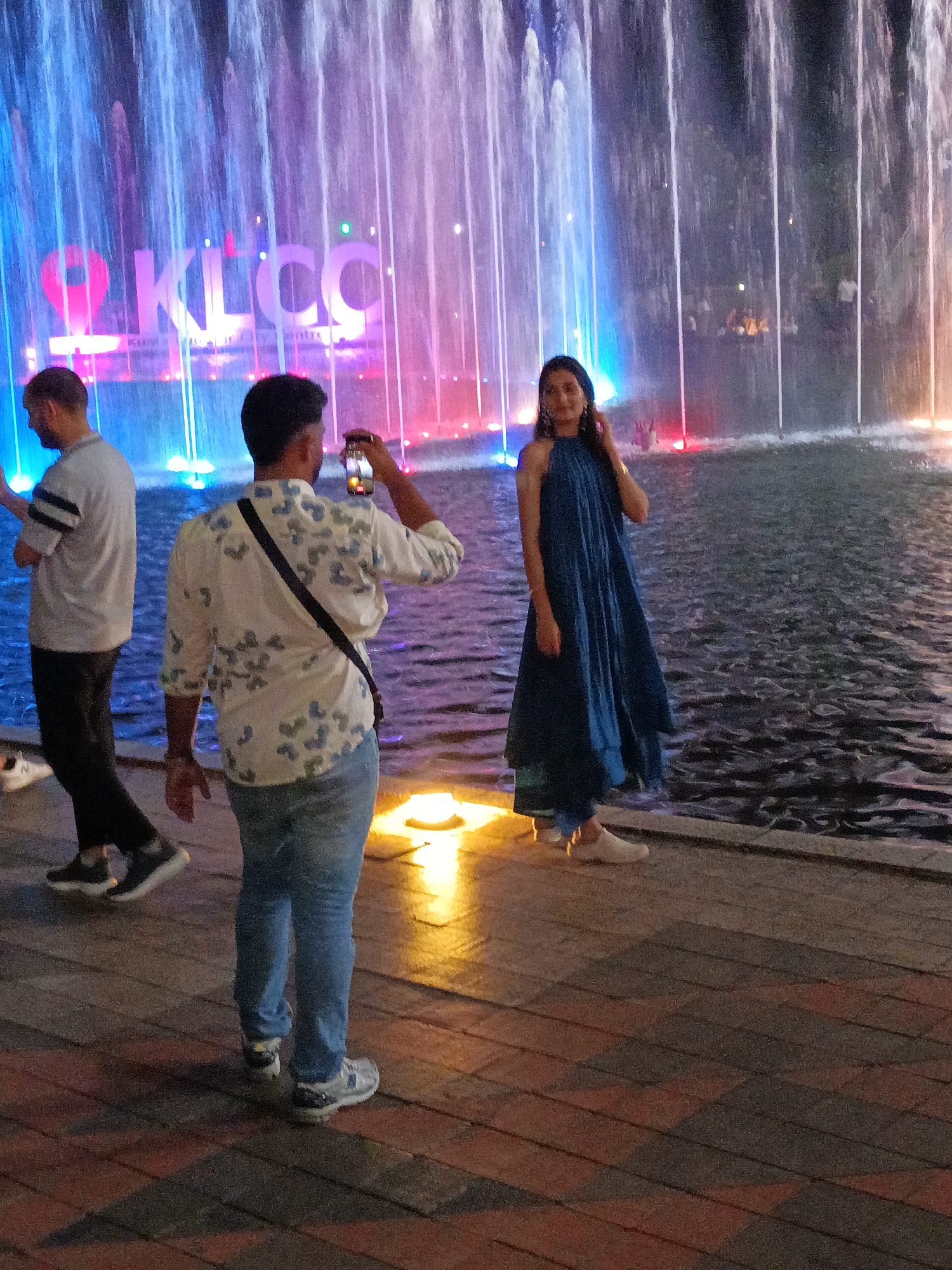 The image captures an outdoor scene at dusk or night, featuring a vibrant water fountain display with colorful lights of blue, pink, and yellow. The fountain is located in front of a prominent sign that reads "KLCC," indicating the location is the Kuala Lumpur City Centre in Malaysia. In the foreground, three individuals are present. On the left, a man with short hair wearing a light-colored shirt with grey stripes on the sleeves and dark pants is walking away from the camera. In the middle, another man is holding up a smartphone to take a photograph of the fountain, his back to the camera. He is wearing a white shirt with a blue floral pattern and blue jeans. To the right, a woman in a long, dark blue sleeveless dress is posing and looking towards the camera, with her right hand touching her hair. She is wearing light-colored slip-on shoes. The ground is paved with interlocking bricks, and the water from the fountain creates a shimmering surface. The overall atmosphere suggests a tourist or recreational area.