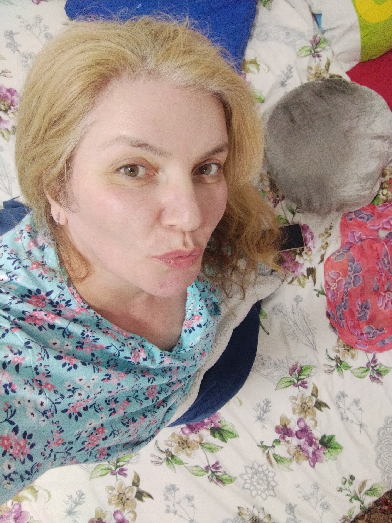 A middle-aged woman with blonde hair and light-colored eyes is taking a selfie. She is wearing a light blue top with a floral pattern and has her lips puckered. She appears to be lying down on a bed with a floral patterned comforter. There are pillows in the background, one is gray and round, another is red and a corner of a green pillow is visible. A red scarf with a floral pattern is also visible on the bed. The image is taken from a close-up, high-angle perspective. There is no information to suggest the location in Al Mas'oudi, United Arab Emirates.