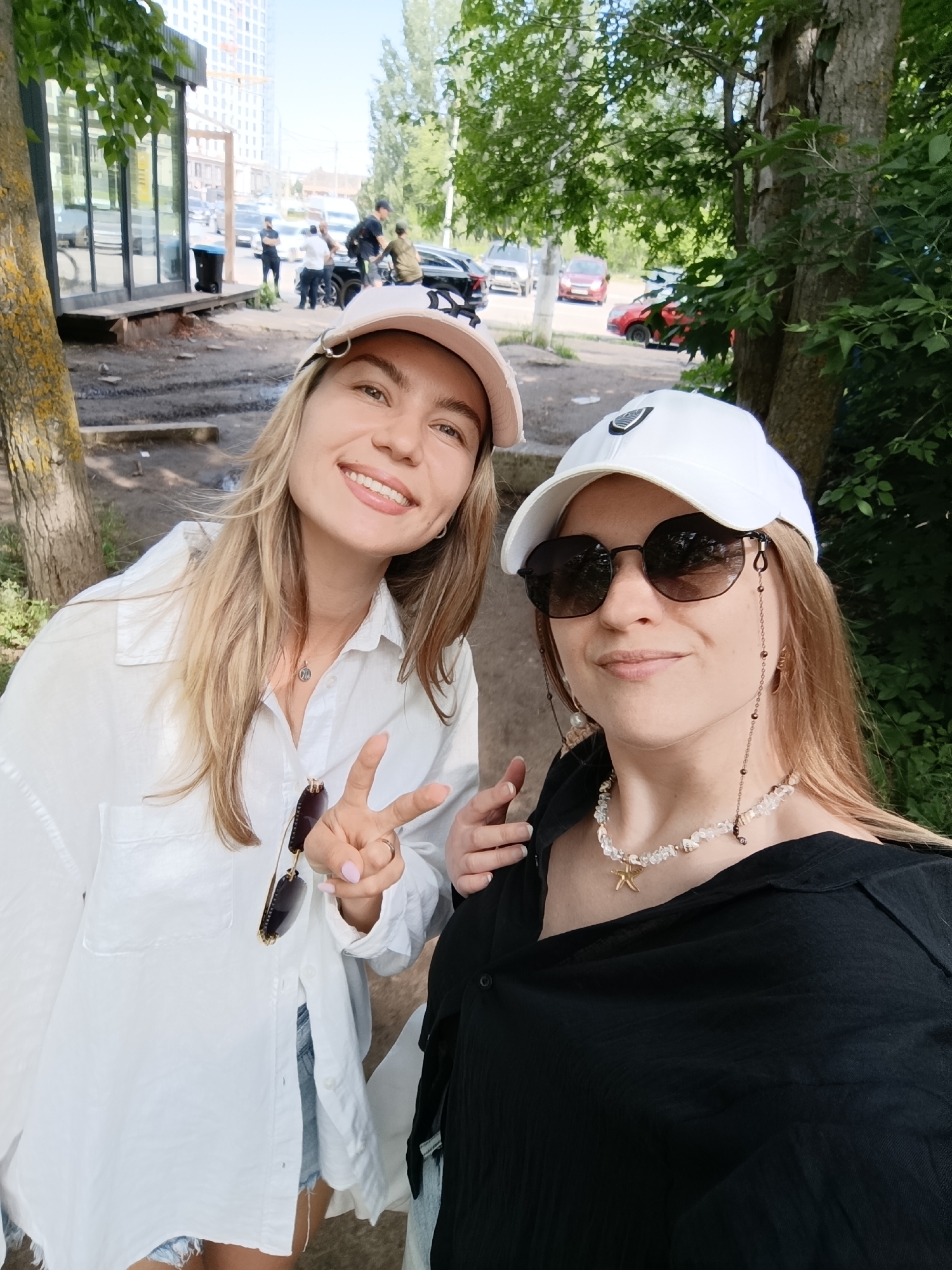 Two women are posing for a selfie in front of a street with cars and trees in the background. The woman on the left is wearing a white button-down shirt, denim shorts, and a light pink baseball cap. She has blonde hair and is smiling, holding up two fingers in a peace sign. She is wearing a necklace and a ring. The woman on the right is wearing a black shirt, sunglasses, and a white baseball cap. She has blonde hair and is also smiling, wearing a necklace with a starfish pendant. She has sunglasses with a beaded chain. The environment appears to be an urban setting with buildings and vehicles visible in the distance.
