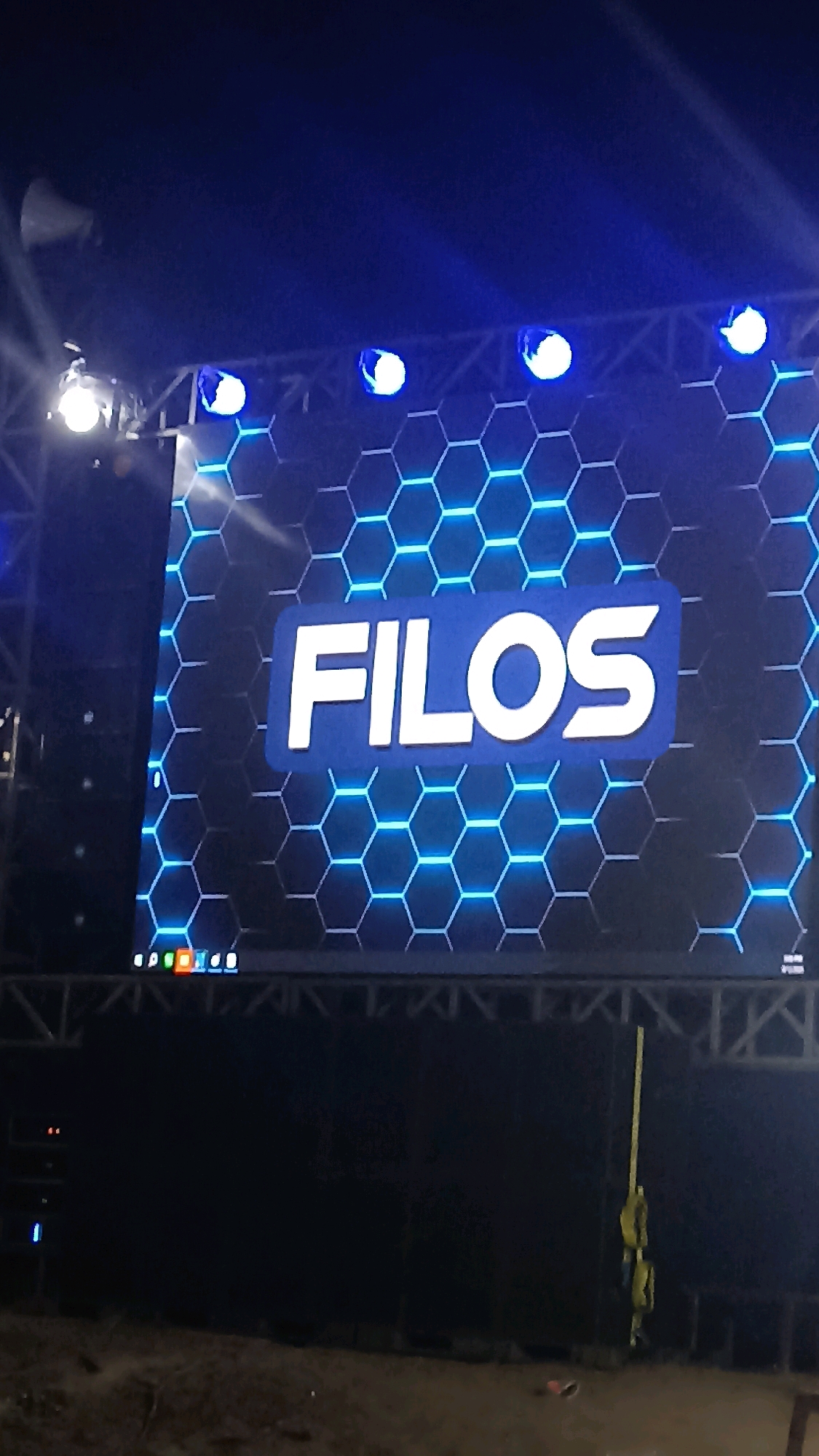 The image shows a large LED screen displaying the word "FILOS" in white letters against a dark blue background with a hexagonal pattern of blue lights. Above the screen, several spotlights are visible, some casting bright white light and others emitting blue light. The screen and spotlights are suspended from a metal truss structure, suggesting an outdoor event or a stage setup. In the lower part of the image, a dark surface is visible, possibly the ground or a stage floor. The overall scene is dimly lit, with the bright lights of the screen and spotlights being the primary sources of illumination. The context of Pacitan, Indonesia, is not directly inferable from the visual elements of the image itself.