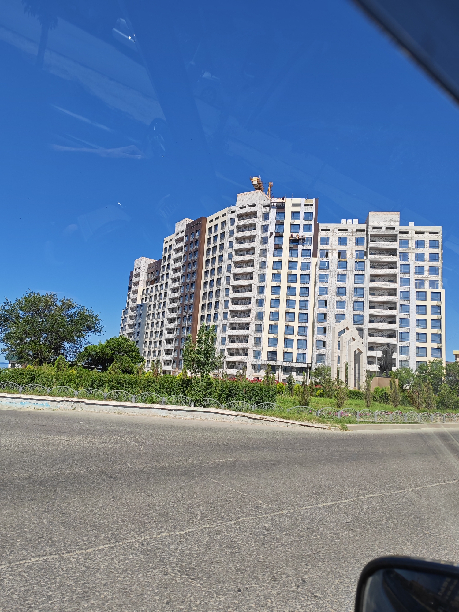 This is an eye-level, wide shot of a large, modern apartment building under construction in Aktau, Kazakhstan. The building is multi-story and has a curved facade with many balconies and windows. A construction crane is visible at the top of the building. In front of the building is a lush green area with trees and bushes, separated from the foreground by a decorative fence and a low wall. A paved road with white lane markings is in the foreground. The sky is clear and blue, with a few white clouds. The image appears to have been taken from a moving vehicle, as there are reflections of a car's interior on the left side of the frame, and a car's side mirror on the bottom right. The overall scene suggests a sunny day in a coastal city.