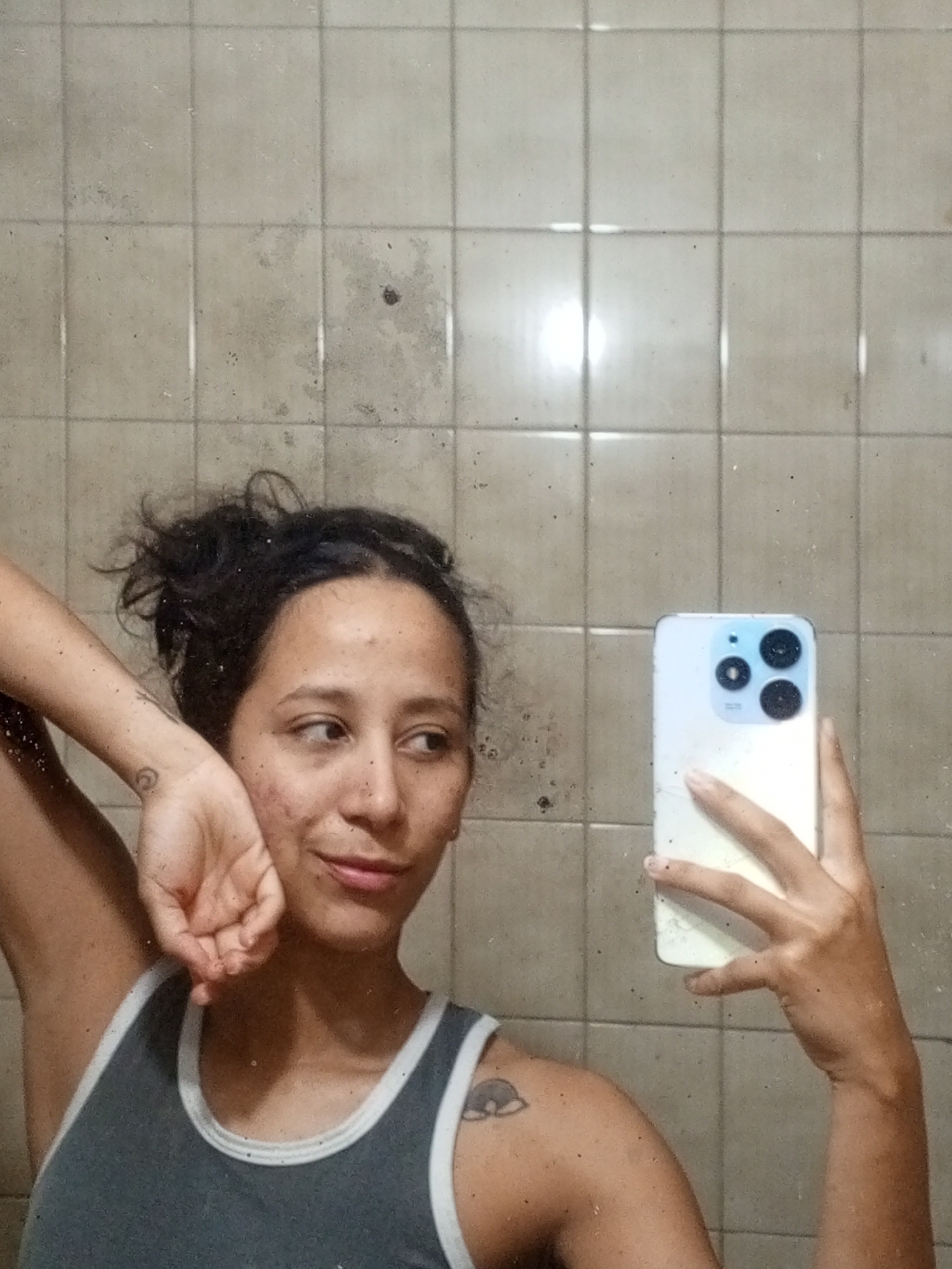 A young woman is captured in a mirror selfie, looking slightly to her right with a soft, enigmatic smile. Her dark, messy hair is pulled up, and her tan complexion shows some visible blemishes. She wears a dark grey tank top with white trim. Her left arm is raised, with her hand gently resting near her face, revealing a spiral-shaped tattoo on her forearm. A stylized eye or cloud tattoo is also visible on her right shoulder. In her right hand, reflected in the mirror, she holds a white smartphone with a distinctive light blue frame around its camera module.
The background is a tiled wall composed of light-colored, possibly off-white or beige, square tiles. The tiles and their grout lines show signs of wear, dirt, and dark discoloration, suggesting an older or less maintained indoor environment. The entire image is overlaid with numerous small, dark speckles, consistent with dust or dirt on the mirror surface where the selfie was taken. This setting is indicative of an indoor space, likely a bathroom or utility area within a home in Porlamar, Venezuela. The woman's expression is calm. There is no discernible text in the image.