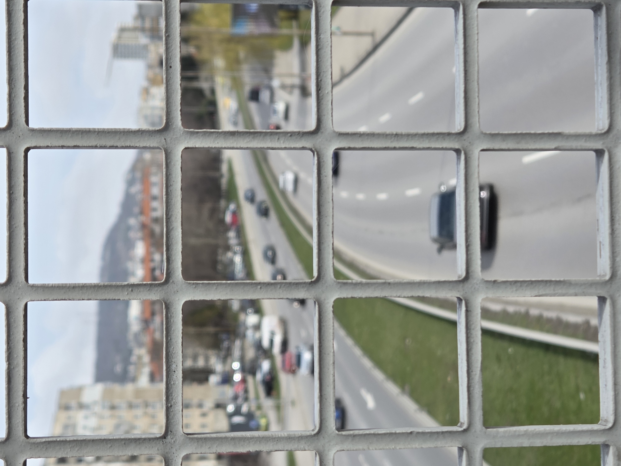 An elevated, matter-of-fact view captures a blurred urban landscape through the sharp, light-grey metal grid of a foreground railing. The grid, composed of horizontal and vertical bars forming uniform square openings, is heavily textured and in sharp focus, dominating the immediate foreground.
Through these square apertures, a multi-lane thoroughfare is visible below, characterized by active traffic. Numerous vehicles, including cars and a white van, are depicted in motion across multiple lanes, marked with white dashed and solid lines. A central green grass median separates opposing directions of traffic.
In the mid-ground and background, the scene is significantly out of focus, revealing various urban elements. Multi-story residential buildings, predominantly light-colored, stand along the road. Distant hills or mountains, partially vegetated, rise against an overcast sky. A construction crane is visible in the far background, indicating ongoing development. The overall environment suggests a bustling urban or suburban area under natural daylight.
This scene is located on a pedestrian overpass above **Vladislav Varnenchik Boulevard (Bul. "Vladislav Varnenchik")** in Varna, Bulgaria.