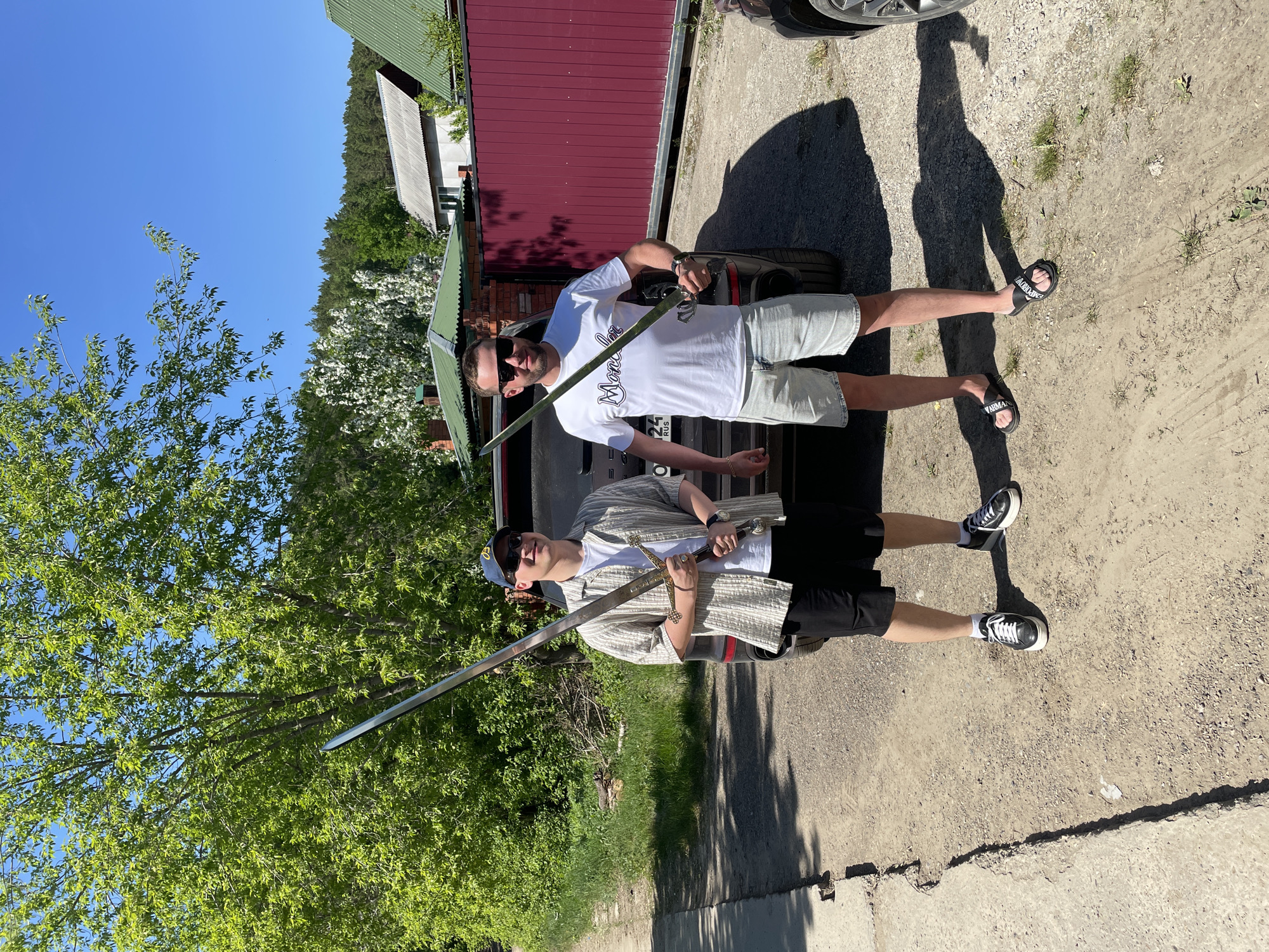 This image, captured in Манский, Russia, depicts two adult males posing outdoors on a clear, sunny day. They are standing on a dirt and gravel surface, which transitions into a cracked concrete strip along the left side of the frame. A dark-colored SUV, possibly black, is positioned between the two men. A portion of its license plate, "025 RUS," is visible behind the man on the right.
The man on the left, appearing younger, wears a dark blue baseball cap with a yellow logo, dark sunglasses, a white t-shirt, an open short-sleeved light-colored striped collared shirt, black shorts, and black and white sneakers with white socks. He holds a large, two-handed metallic sword with an ornate, possibly gold-toned, crossguard and pommel. The blade extends upwards and to the left. He is smiling and looking towards the camera.
The man on the right, appearing slightly older, wears dark rectangular sunglasses, a white short-sleeved t-shirt with a black "Moncler" logo, light blue denim shorts, and black slide sandals with white "ARMANI" branding. He holds a shorter metallic sword with a darker hilt in his right hand, with the blade also extending upwards and to the left. His left hand is resting on his hip. He is also smiling and looking towards the camera.
In the background, behind the subjects and the SUV, there is a red corrugated metal fence or gate. Beyond this fence, several residential structures are visible, including a white building with a grey corrugated roof and another building with a green corrugated roof. A dense line of green coniferous trees forms a forest on the horizon, beneath a clear blue sky. Directly behind the buildings, a large tree with abundant white blossoms is prominent. To the far left and behind the man on the left, lush green deciduous trees with full foliage are visible. The strong sunlight casts distinct shadows of the men and the vehicle onto the ground.