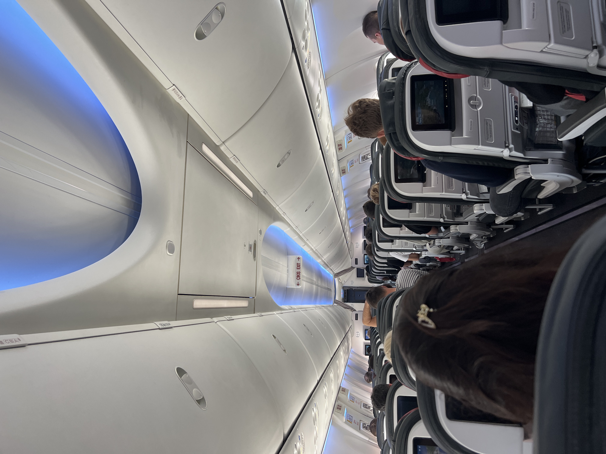 This media file captures the interior of an airplane cabin, likely taken from the aisle looking towards the rear. Numerous passengers are visible, primarily from behind, seated in their airplane seats. The cabin features overhead bins along both sides and individual entertainment screens on the seatbacks in front of each passenger.
The overall scene depicts a typical airplane environment, suggesting passengers are either waiting for departure, in flight, or awaiting disembarkation. The cabin is well-lit with artificial overhead lighting, complemented by blue accent lighting that emanates from the curved panels along the ceiling and walls, possibly indicating mood lighting for an evening or night flight.
Visible text includes seat row/area markers such as "16 FED" and "16 CBA" on the overhead bins. A prominent bilingual sign reading "← ÇIKIŞ EXIT →" is seen further down the aisle, indicating an exit. "ÇIKIŞ" is the Turkish word for Exit, which aligns with the provided location context of Arnavutköy, Turkey, a district often associated with Istanbul Airport. Another "ÇIKIŞ EXIT" sign is also visible, along with "CBA". The image shows a modern aircraft interior with comfortable-looking seats.
