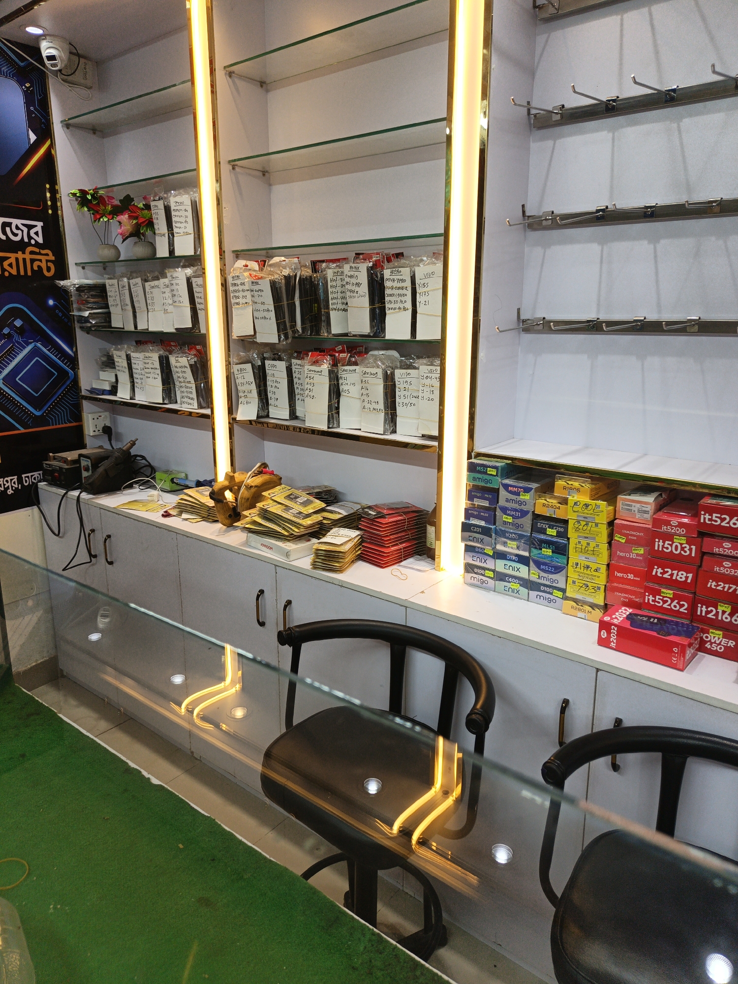 The image depicts the interior of a mobile phone accessories shop in Dhaka, Bangladesh. The shop is well-organized, with shelves stocked with various mobile phone accessories, including screen protectors, cases, and chargers. The lighting is bright, with a prominent vertical LED strip illuminating the shelves. The shelves on the left are filled with packaged screen protectors, many with handwritten labels indicating phone models. The shelves on the right display boxed mobile phone accessories. In the foreground, a glass counter reflects the shop's interior. Two black chairs with curved backs are visible, suggesting a seating area for customers. A green carpet covers the floor in front of the counter. The overall scene conveys a commercial environment typical of electronics retail in the city.