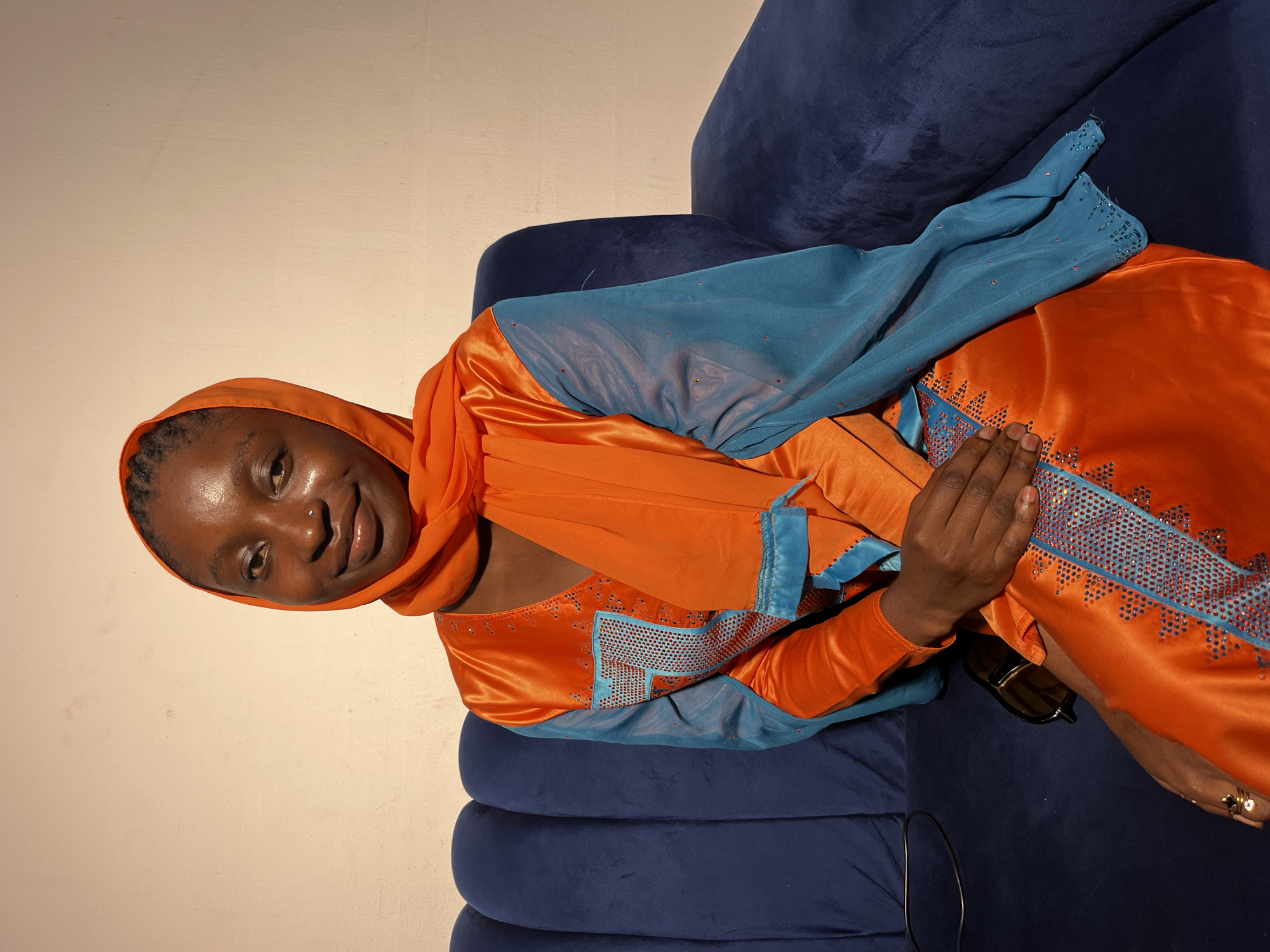 A young Black woman with braided hair is shown from the chest up, wearing an orange headscarf and a shimmering orange and teal dress adorned with rhinestones. She is seated on a dark blue velvet couch, with her hands clasped in her lap. Sunglasses are visible on the couch next to her. The background is a plain, light-colored wall. The lighting suggests an indoor setting, possibly a home or studio.
