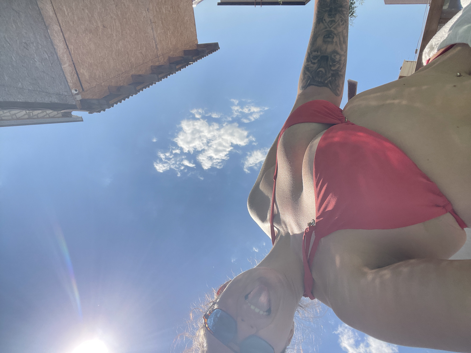 A woman is wearing sunglasses and a red bikini. She has a tattoo on her arm. She is outdoors, with a blue sky and clouds in the background. Part of a building structure is visible above. The location is Мосметаллоконструкция Онт, Russia.