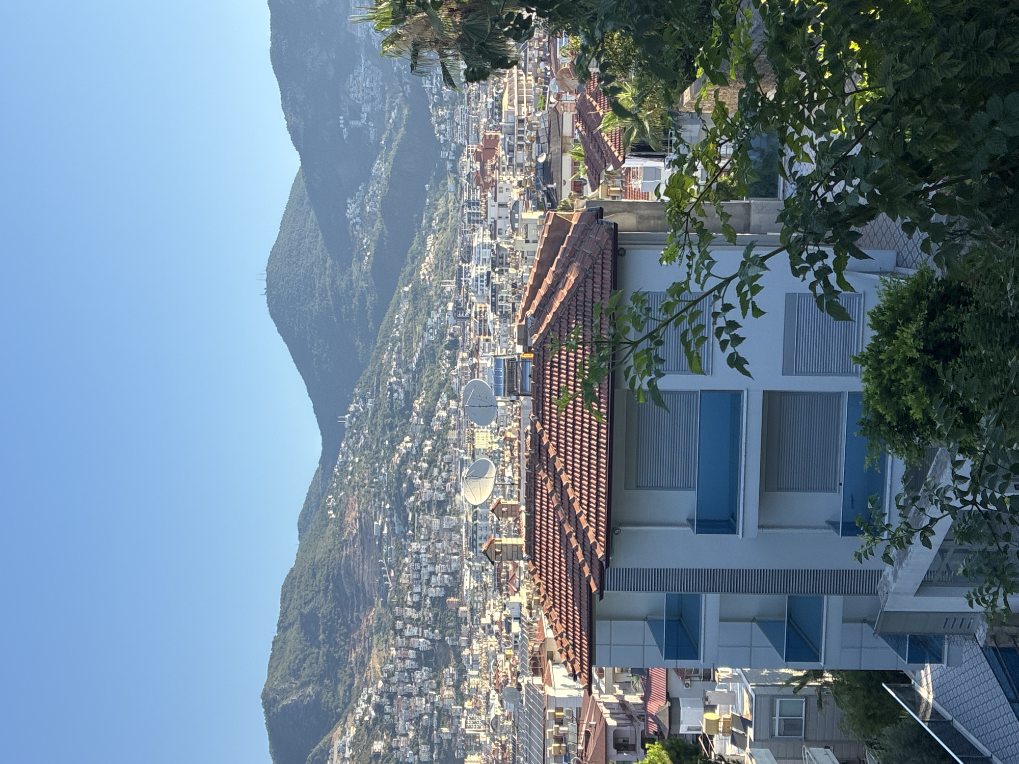 The image captures a panoramic view of Alanya, Turkey, on a clear day. The foreground features the roof of a building with red tiles, and a portion of a white multi-story building with blue balconies. In the mid-ground, a dense urban landscape unfolds, characterized by numerous buildings of varying heights and architectural styles, many with similar red-tiled roofs. Satellite dishes are visible on several rooftops. The background is dominated by a chain of lush, green mountains, dotted with white buildings that extend up their slopes. The sky is a bright, cloudless blue, indicating fair weather. No people are visible in the scene. The overall impression is one of a coastal city nestled against a mountainous backdrop.