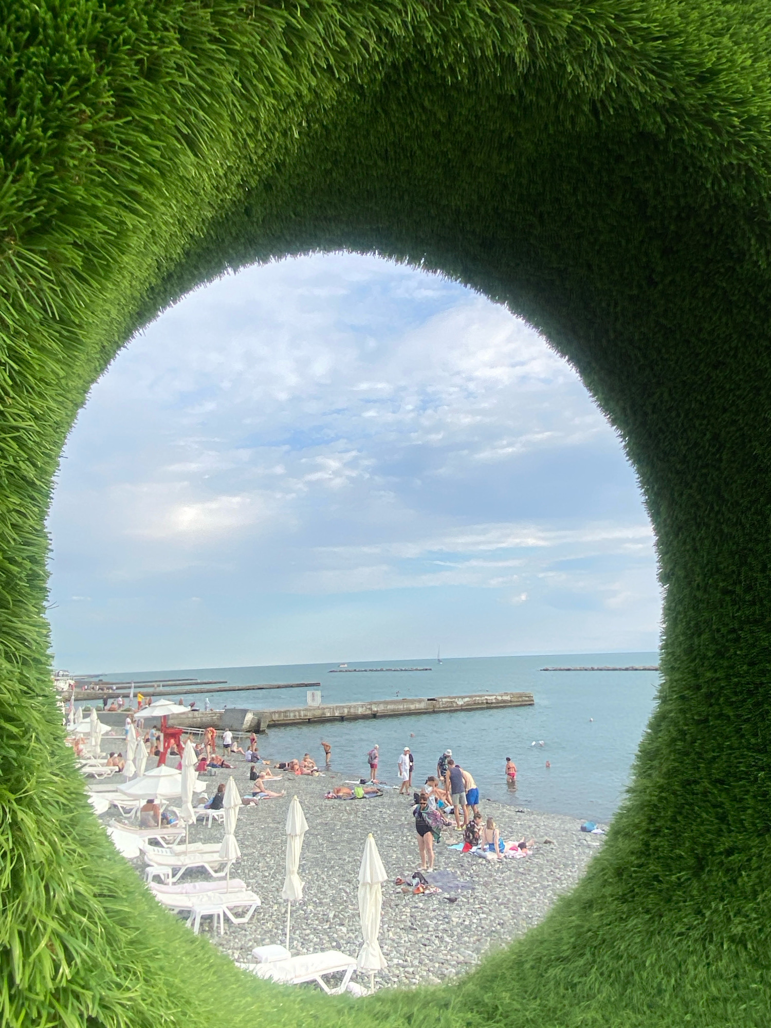 The image depicts a beach scene in Sochi, Russia, framed by a large, circular opening covered in artificial green grass. The foreground is dominated by this grass, creating a naturalistic tunnel effect. Beyond the opening, a pebble beach stretches towards the sea. Numerous people are visible on the beach, some sunbathing on lounge chairs, others walking, and some in the water. Beach umbrellas and lounge chairs are set up on the shore. The sea is calm, with piers extending into it, and several boats can be seen in the distance. The sky is partly cloudy, suggesting a pleasant day. The overall atmosphere is one of a relaxed, summer vacation.