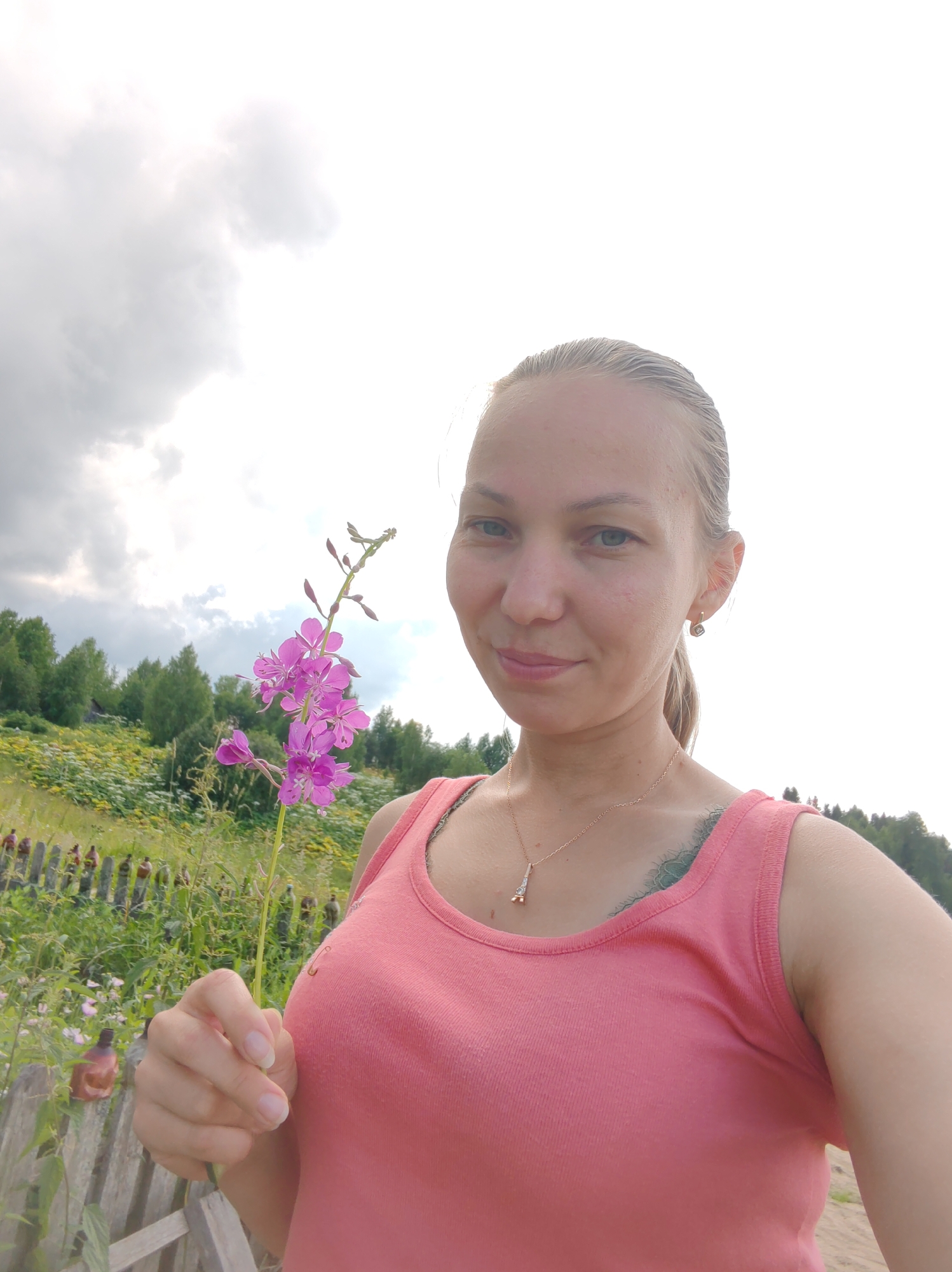 In Чухлэм, Russia, a young woman with blonde hair pulled back in a ponytail stands outdoors, holding a single pink wildflower. She is wearing a coral-pink tank top and a delicate gold necklace with a pendant. A light tattoo is visible on her chest, partially obscured by her shirt. The background features a wooden fence, lush green vegetation, and a wooded hill under a cloudy sky. The overall impression is a serene, natural setting on a summer day.