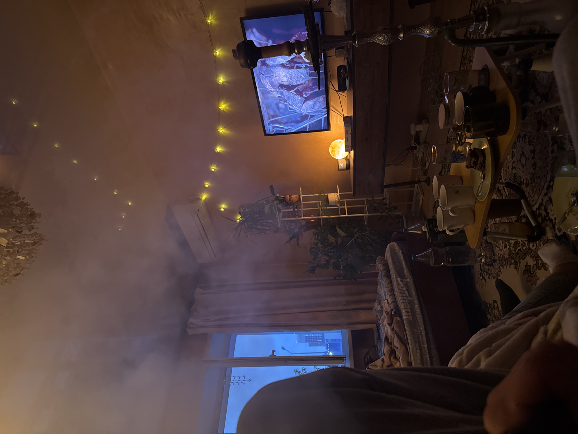The scene is an interior shot of a room, identified as being in the city of Perm, Russia. The time appears to be evening or night, indicated by the dark-blue exterior visible through the window and the reliance on artificial lighting. One individual is partially visible in the foreground, seated on the floor or a low surface with their back to the viewer. They are wearing a light-colored, possibly hooded, garment over another layer, dark trousers, and light-colored socks. Their right arm is visible in the lower left of the frame. The room is illuminated by multiple artificial light sources: a partially visible overhead chandelier, two strands of yellow fairy lights (one strung across the ceiling and another above the television), and a spherical, warm-toned lamp on a media console. The television screen emits a bluish light. A significant volume of diffused smoke or vapor is present in the upper half of the room. To the left, a large, multi-pane window reveals a dark urban environment, including a street light and distant buildings. Light-colored curtains are drawn over part of the window. A decorative hanging object is visible near the top left of the window frame. Below the window, a mattress resting on a low bed frame is covered with a patterned blanket. The word "Bonheur" is visible repeatedly on the mattress fabric. Against the back wall, a white, multi-tiered metal shelving unit holds several potted green plants. One pot on the shelf features a painted face. More potted plants are positioned on the floor nearby. A white split-system air conditioning unit is mounted high on the wall to the right. A flat-screen television is mounted on the wall, displaying an image of textured, dry leaves or organic material, predominantly in blue and brown tones. Below the television, a dark wooden media console supports a spherical lamp, a small digital clock displaying "26", and a white rectangular electronic device. Electrical cables and an outlet strip are visible on the floor behind the console. In the right foreground, an ornate, silver-colored hookah with a dark top is prominently positioned, partially obscuring the television. A low, rectangular, yellow table with foldable legs is situated in the foreground. On this table, a white plate holds three pieces of dark baked goods (resembling brownies) and a green fork. Additionally, four mugs (two white, one dark, one clear) and two clear drinking glasses are on