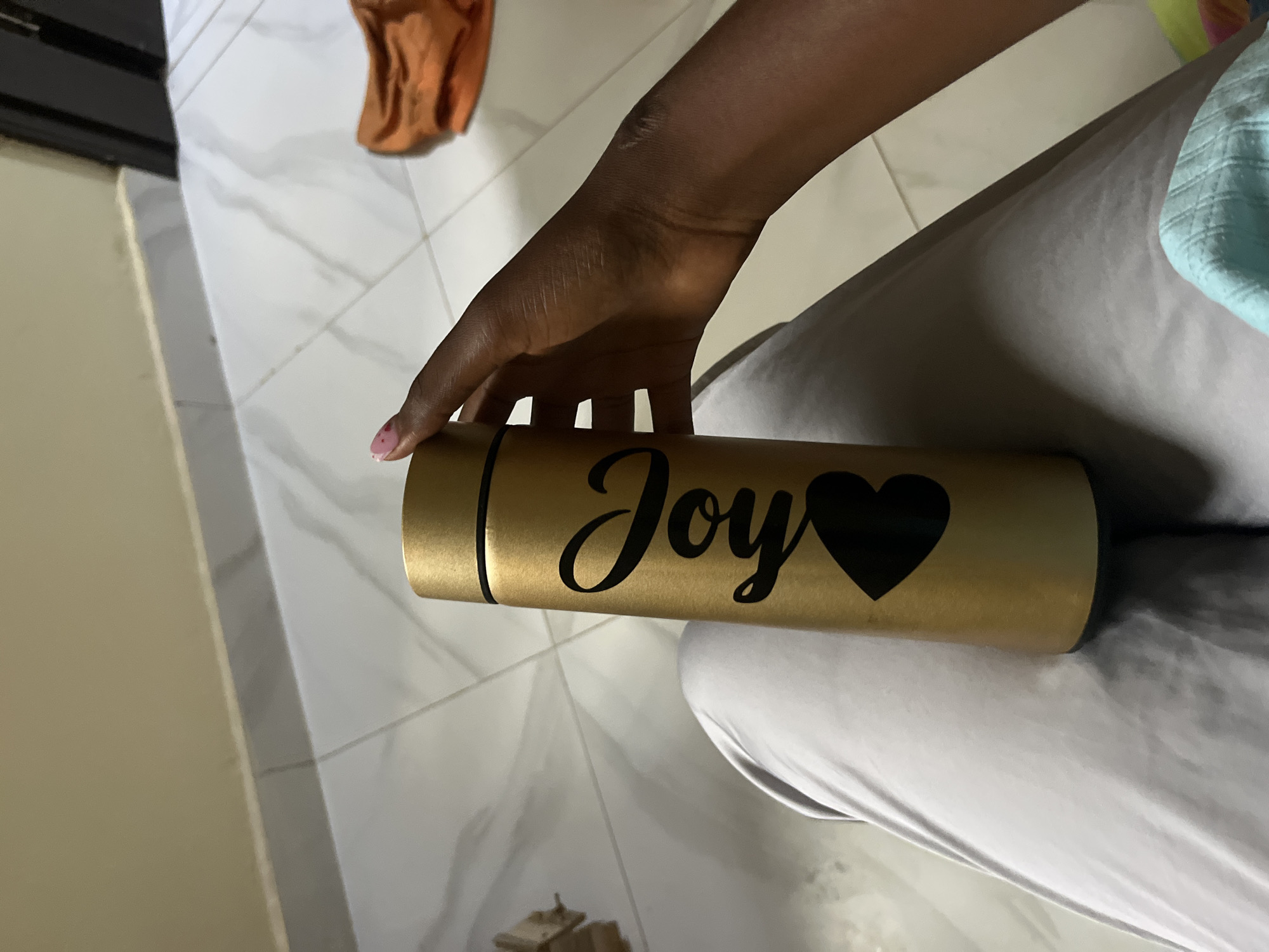 A person's hand, with a pink-tipped fingernail, holds a gold-colored metal water bottle. The bottle has "Joy" written in black script on its side, with a black heart beneath the word. The hand and bottle are positioned over a person's lap, covered by light gray fabric. The background shows a tiled floor and a beige wall.