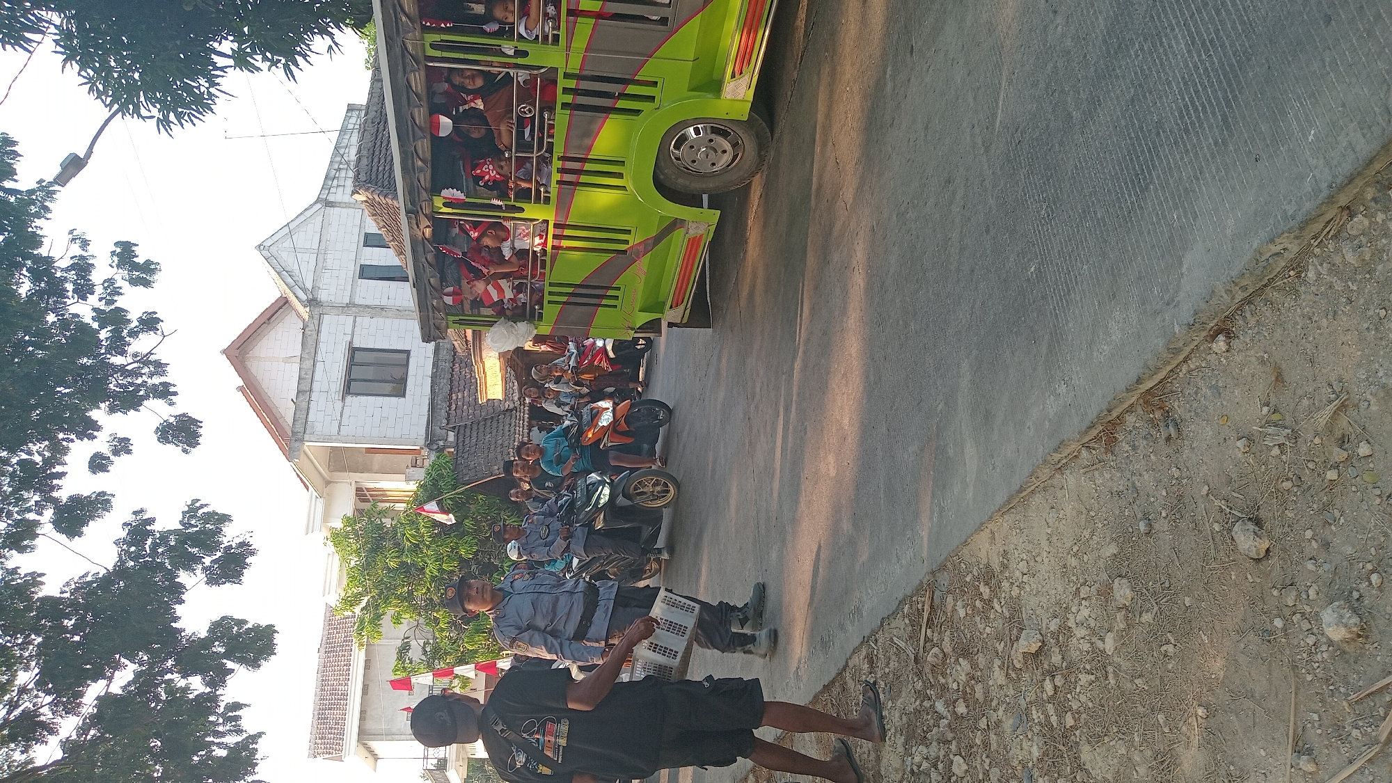 In Kedungwaru, Indonesia, a group of people are gathered on a street. In the foreground, a person wearing a black t-shirt and shorts stands facing away from the camera, holding a white object. To their left, a man in a blue uniform stands on a motorcycle, looking towards the viewer. Several other motorcycles are parked or moving on the street. On the right side of the image, a green open-sided vehicle is filled with passengers, many of whom are holding small Indonesian flags. The background shows several houses and trees, with a bright sky overhead. The overall scene appears to be a street celebration or parade.