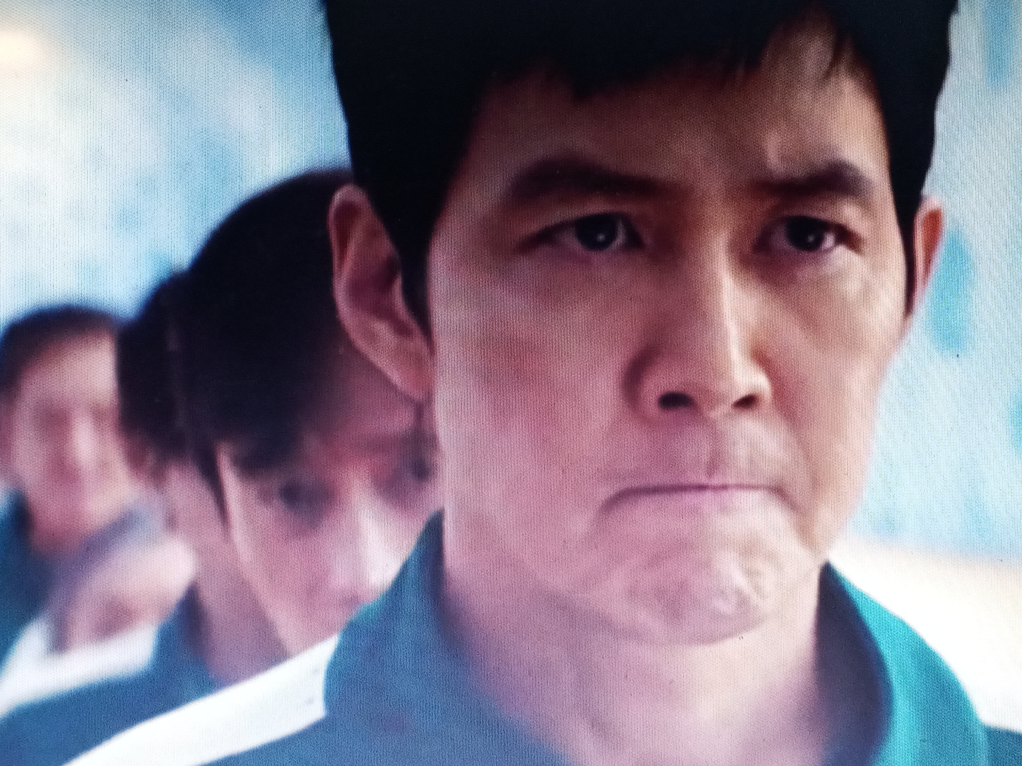 A medium-close-up shot shows an Asian man in a blue uniform, standing in a line with other people who are slightly blurred in the background. He has short, dark hair and a serious expression on his face. The background appears to be a light blue or white, possibly a wall or other surface.