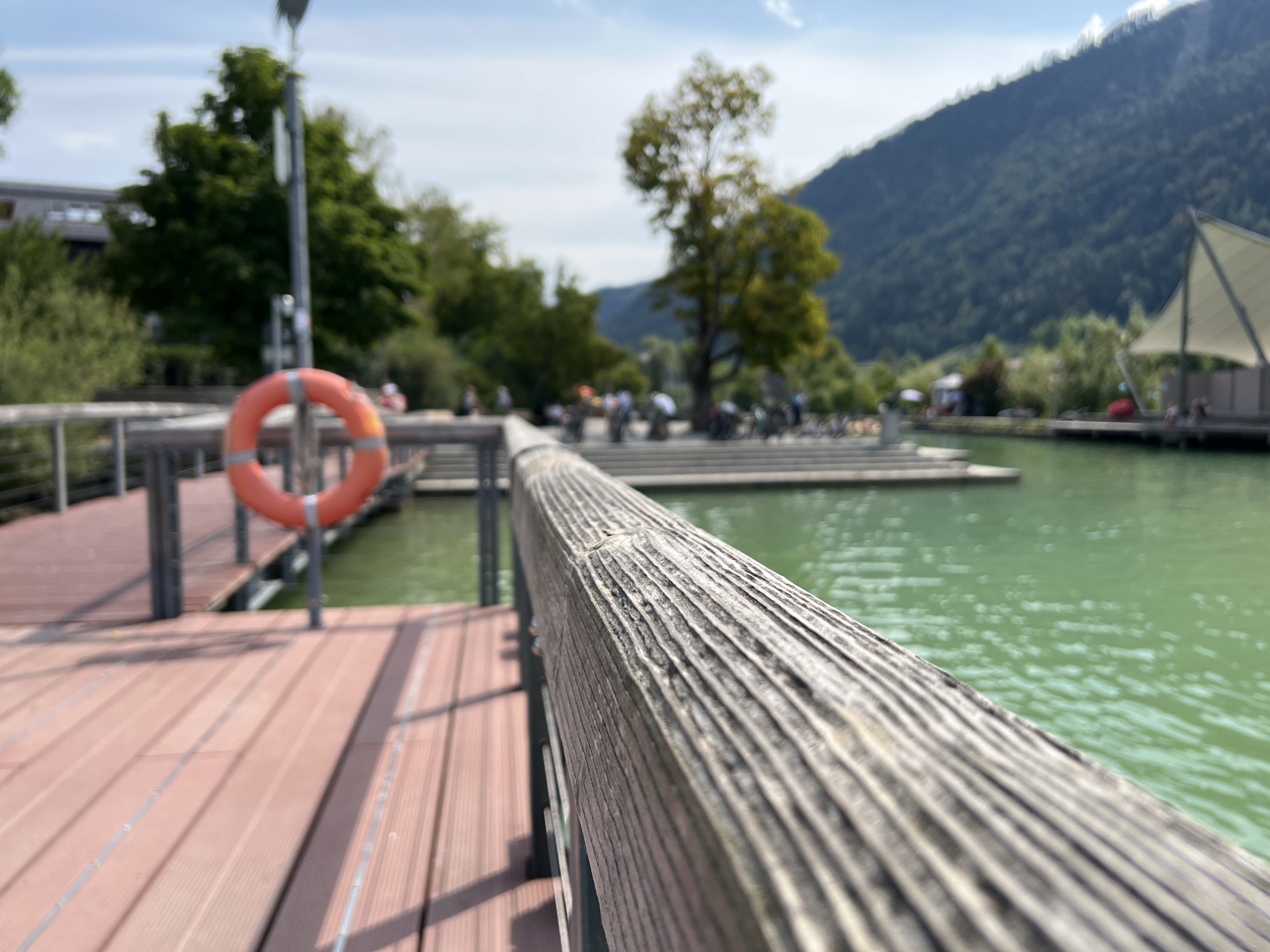 This image depicts a scene from the area around the Großer Alpsee lake in Immenstadt im Allgäu, Germany. In the foreground, a wooden railing of a pier is prominently featured, showing detailed wood grain. To the left, a red life preserver hangs on a metal post, positioned over a wooden pier made of reddish-brown planks. Beyond the pier, the bright green water of the lake is visible, with subtle ripples on its surface. In the middle ground, a paved area with several bicycles suggests a recreational zone. Further back, lush green trees and vegetation line the shore, and a building with a light-colored facade is partially visible. In the background, a steep, tree-covered mountain slopes down towards the lake, under a partially cloudy sky. The overall scene conveys a peaceful, outdoor environment, likely a public area for recreation and leisure by the lake.