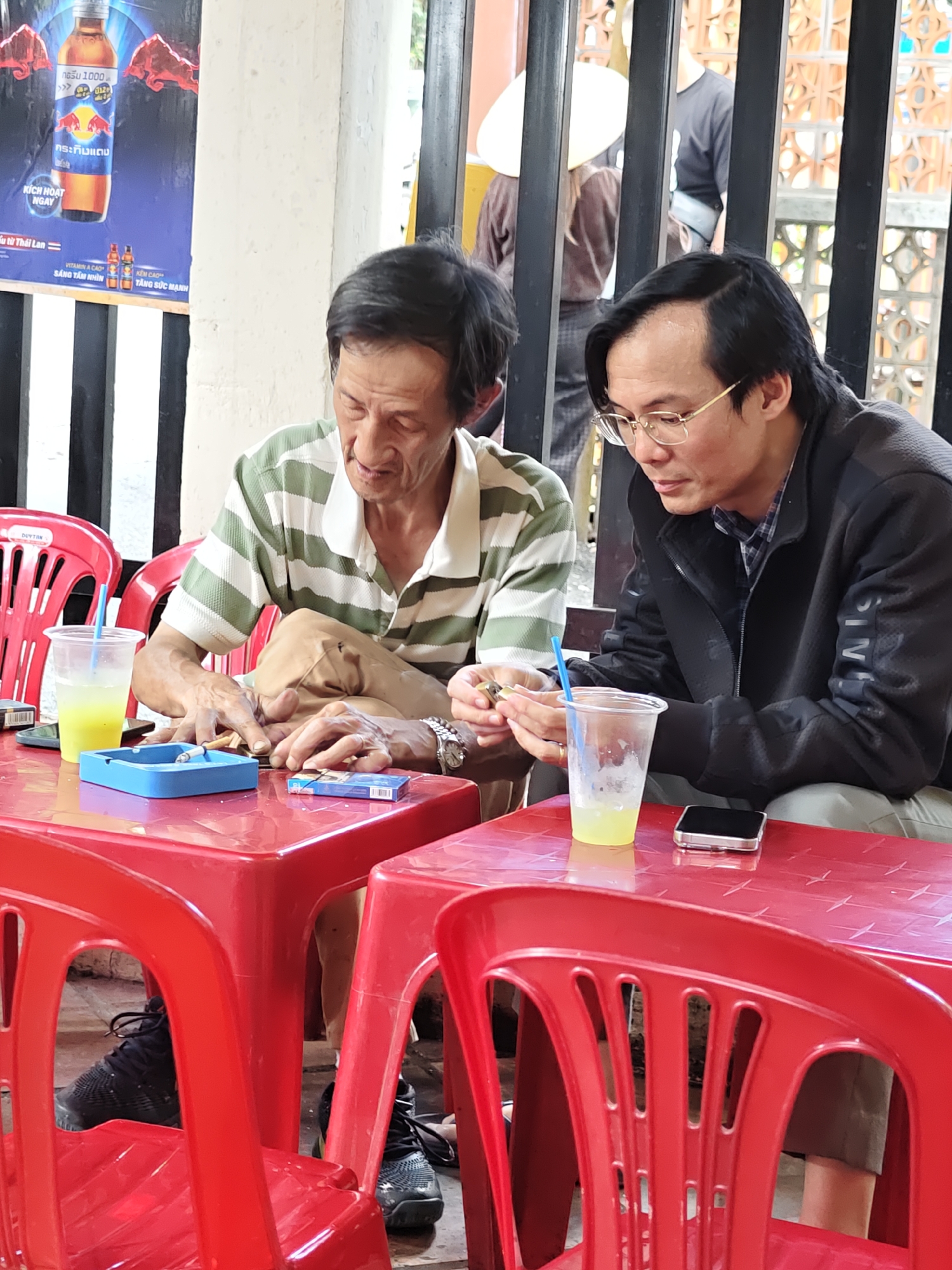 Two Vietnamese men are seated at a red plastic table, likely at an outdoor eatery or cafe in Da Lat, Vietnam. The man on the left, with thinning dark hair and a striped green and white polo shirt, is leaning forward, looking down at his hands as he handles a cigarette. He has visible veins on his hands and is wearing a watch. In front of him, on the table, is a blue ashtray with a lit cigarette in it, and a pack of cigarettes. The man on the right, wearing glasses and a dark jacket over a plaid shirt, is also looking down at the table, holding a lighter and a cigarette. A clear plastic cup with a yellow beverage and a straw is positioned in front of him, as is a smartphone. Red plastic chairs surround the table, and in the background, a black metal fence with an ornate pattern is visible, along with other patrons. A poster of a Red Bull energy drink is also visible on a wall behind the man on the left. The environment appears to be casual and relaxed.