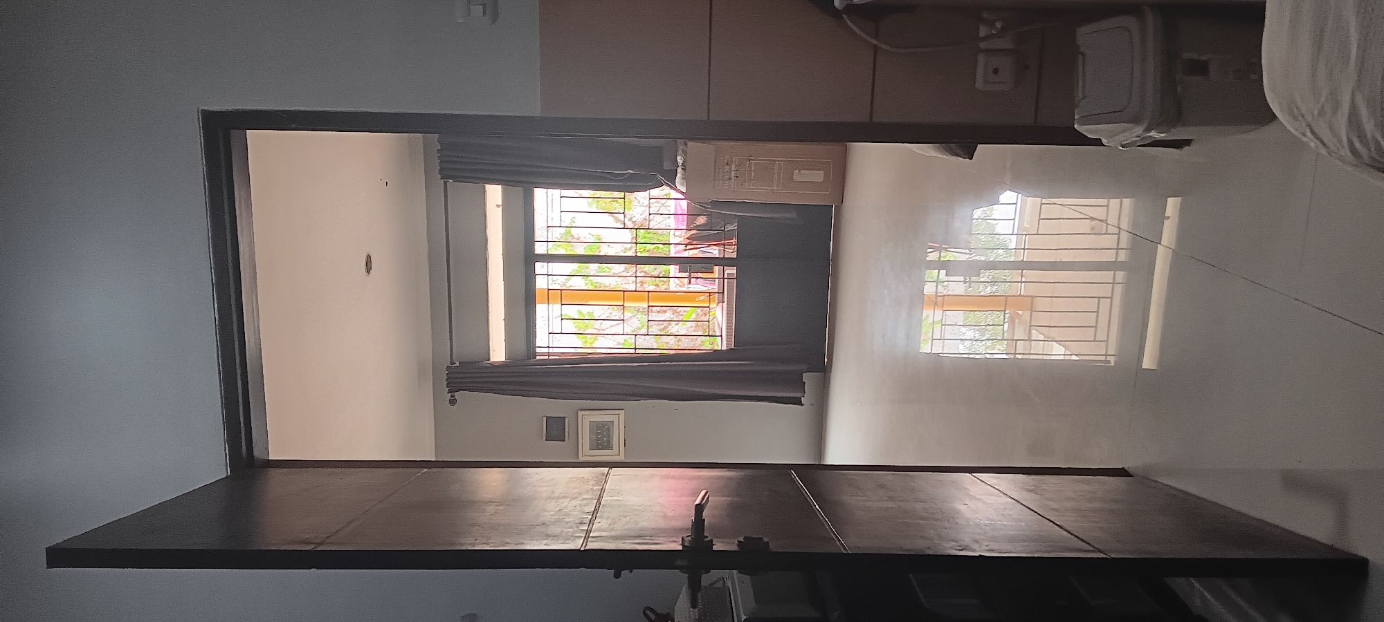 The image shows a view from a doorway looking into a room. The room has a tiled floor that reflects the light. To the left is a dark wooden door, partially ajar, revealing its inner surface. On the right wall, a power outlet is visible, and next to it is a gray plastic bin with a lid. Straight ahead, a large window with a metal grate covers the lower half, and curtains frame the upper half. Outside the window, greenery and part of a railing are visible. The ceiling appears to be white with a single circular light fixture. The scene is indoors, likely in a residential setting in Depok, Indonesia.