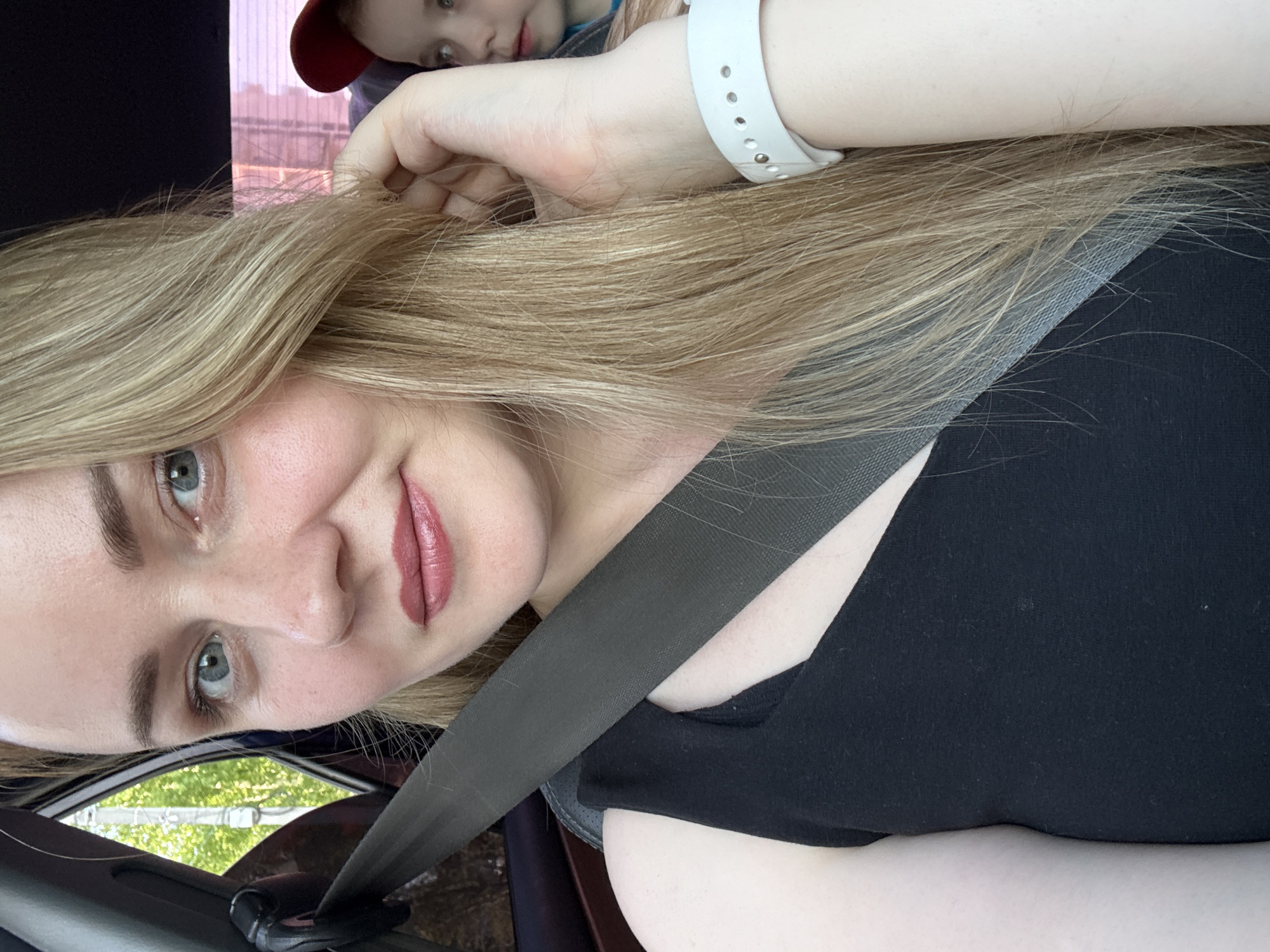 A woman with long blonde hair, wearing a black shirt and a seatbelt, is taking a selfie in a car. The image captures her face, including her blue eyes, pink lips, and fair skin. A child with a red baseball cap is partially visible in the background, behind the woman. The location is identified by a glimpse of buildings outside the car window as Chaikovsky, Russia.