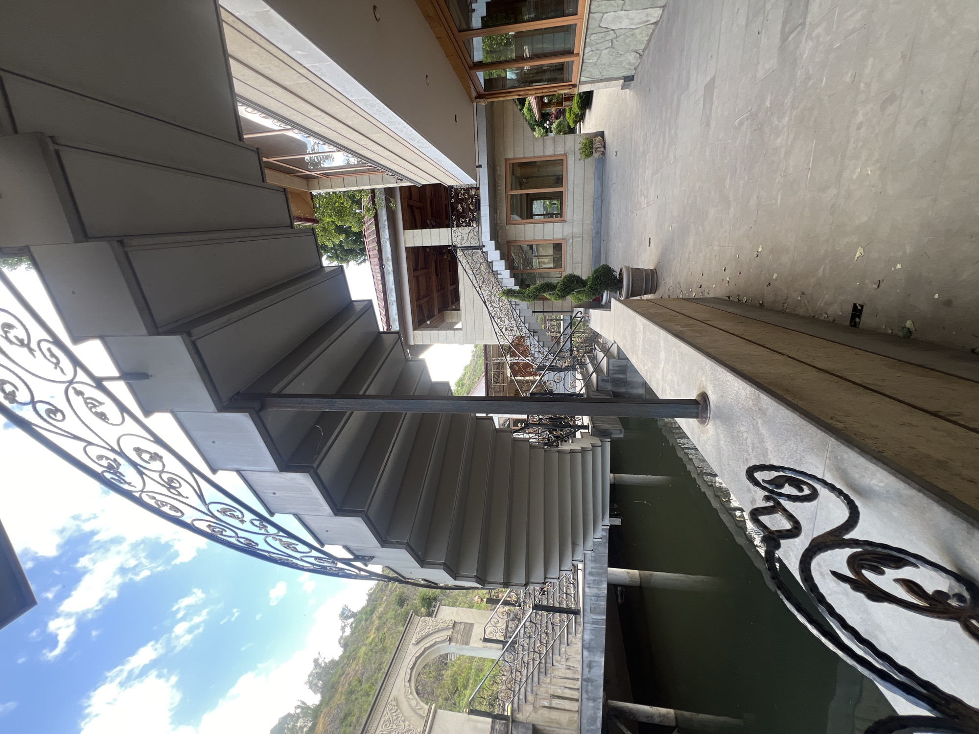 The image captures an outdoor architectural complex in Yerevan, Armenia, seen during a bright, sunny daytime with a clear blue sky and scattered white clouds. The perspective is from a lower vantage point, looking across and upwards at the various structures.
In the foreground, a prominent set of concrete or stone stairs with ornate black wrought iron railings ascends from the lower left towards the center of the frame. These railings feature intricate scrollwork. The stairs appear to connect different levels of the complex.
To the right and further back, several buildings are visible, characterized by stone or stucco facades and rectangular windows with wooden frames. One building in the mid-ground has a reddish-tiled roof and what appears to be an open-air seating area with decorative ceilings. Pathways and additional stairs with similar wrought iron railings lead around and between these structures. Potted topiary plants are strategically placed along the walkways and near the buildings, adding greenery to the scene.
Beneath some of the walkways and structures, particularly in the lower right, a tranquil body of green-hued water, likely a canal or narrow river, flows. Support pillars are visible within the water, upholding the overlying paths. In the distant background, rolling hills or mountains covered with vegetation rise under the bright sky, and a large, decorative stone archway is partially visible on the hillside.
There are no people or specific activities taking place, rendering the scene peaceful and still. No visible text is present within the image.
