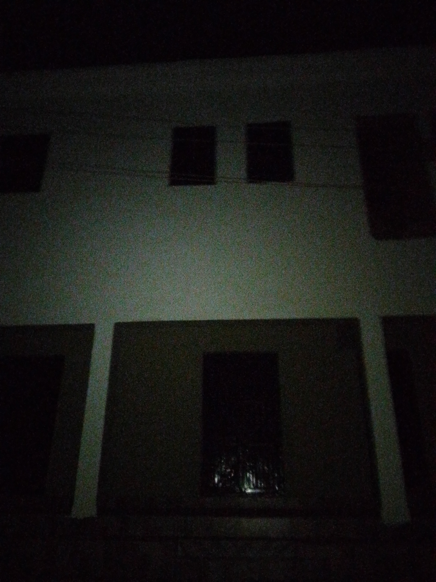 The image shows a dimly lit, two-story building at night. The building appears to be made of a light-colored material, possibly concrete or stucco. The upper level has three dark, rectangular openings, likely windows or doorways. A horizontal line runs across the upper level, possibly an electrical wire. The lower level also has dark openings. The picture was taken in Barikin Gidar Kanat, Nigeria.