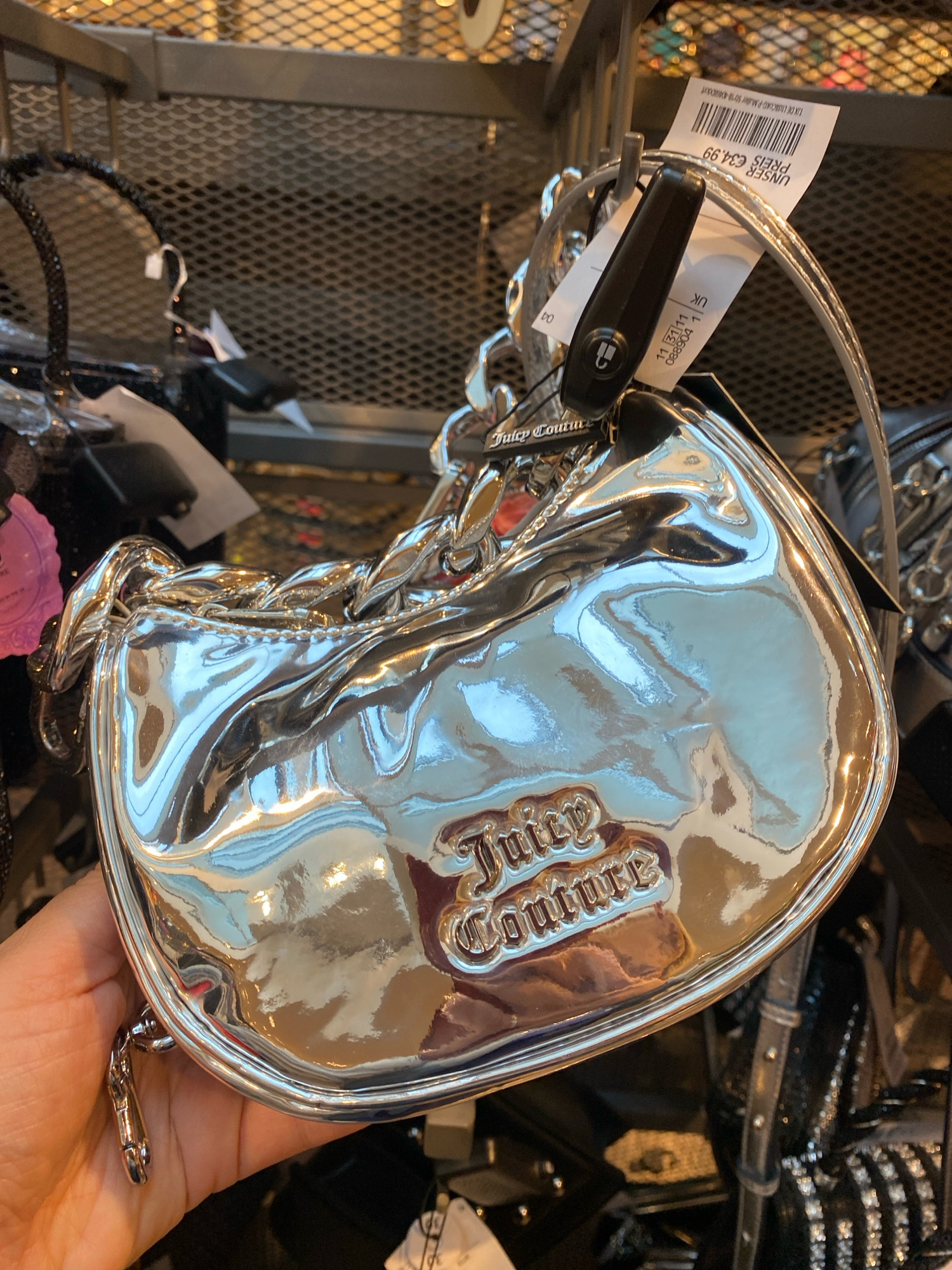 A shiny, metallic silver Juicy Couture handbag with a quilted texture is being held by a person's hand, displaying the brand's logo in gold lettering. The handbag features a chain strap and a black security tag. In the background, other bags and store shelving are visible, suggesting a retail environment. A price tag is attached to the handbag, indicating "UNSER PREIS €34.99" and "UK". While the image does not explicitly state the location, the currency and price format suggest it might be a store in Vienna, Austria.