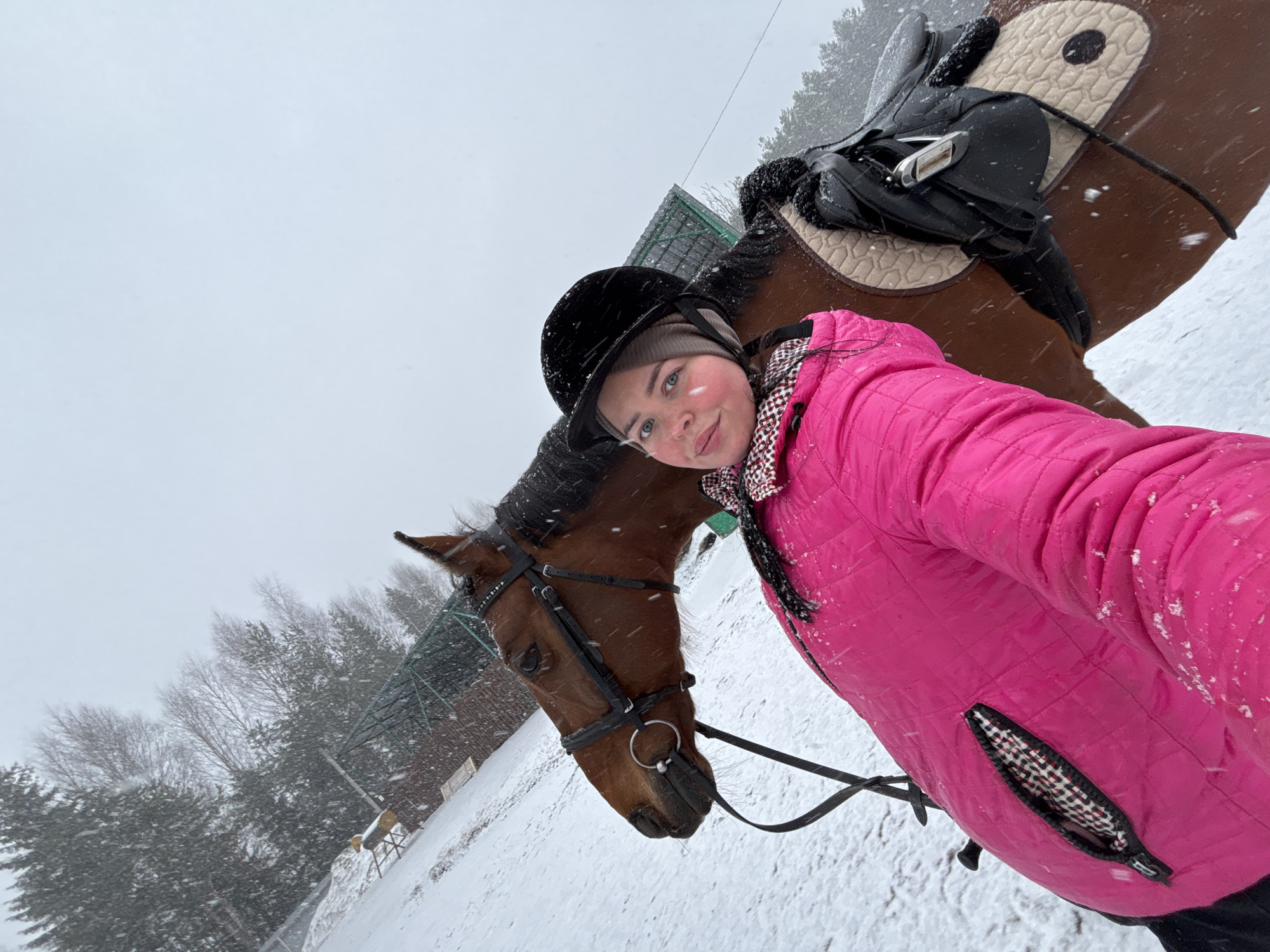 A selfie image shows a young woman standing next to a brown horse in a snowy outdoor environment. The woman is wearing a black riding helmet, a beige knit cap, and a bright pink quilted jacket. She is smiling at the camera. The horse is wearing a bridle and saddle. Snow is falling. Trees are visible in the background, and a building with a green roof is also visible. Based on visual cues, it is likely that this image was taken in a rural or suburban setting. The given information includes the city of Русский Вожой, Russia.
